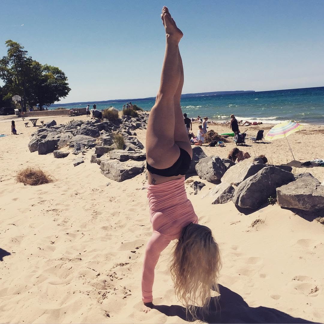 Person performing a handstand on sandy beach with rocks, ocean, and people in background.