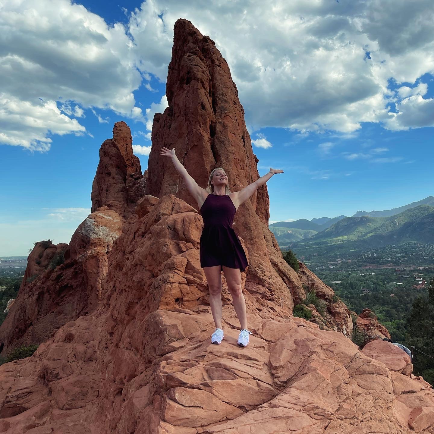 Person standing on rocky terrain with arms raised, large rock formation behind, blue sky with clouds, distant mountains.