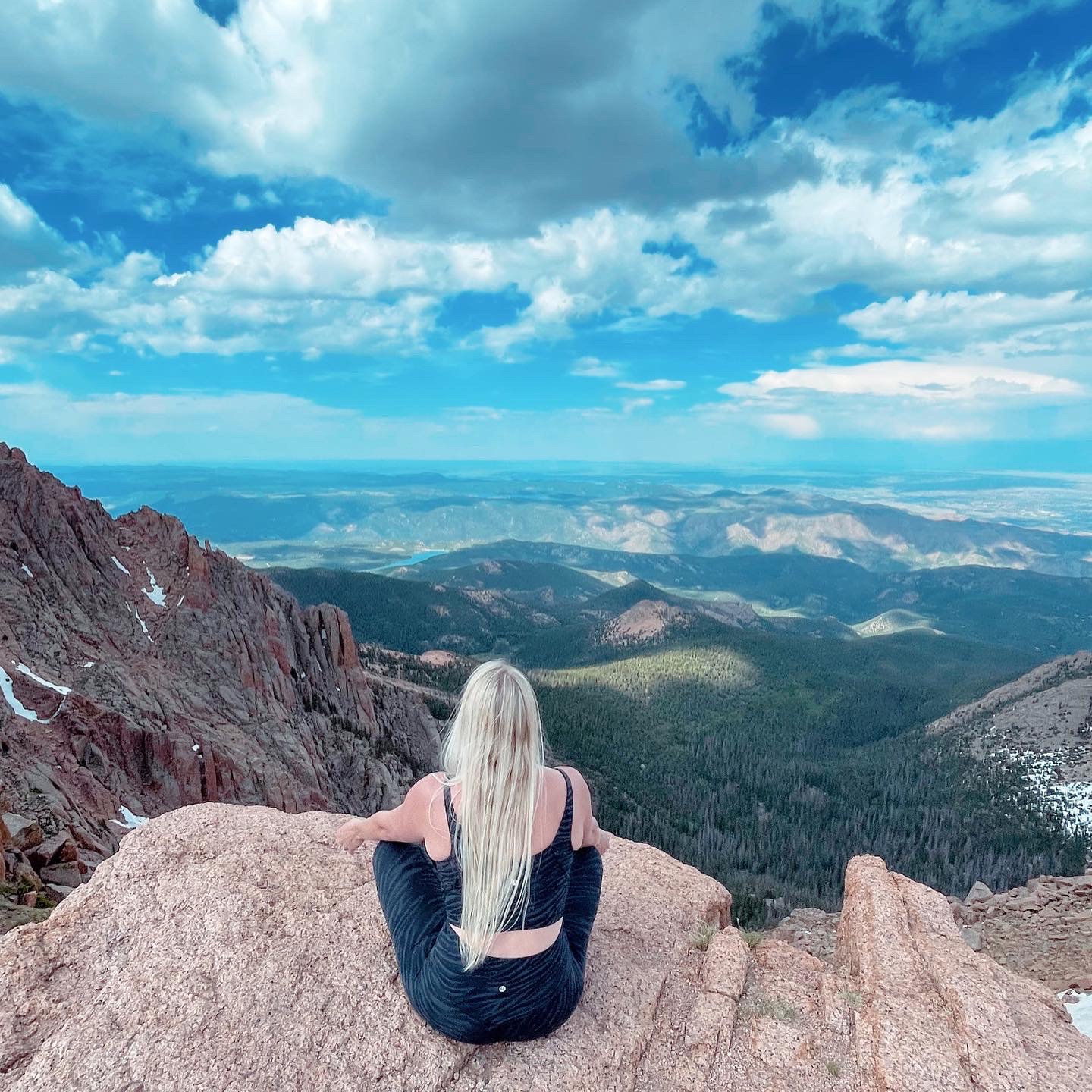 Person with long blonde hair sitting on rocks overlooking a mountain landscape with blue sky and clouds.