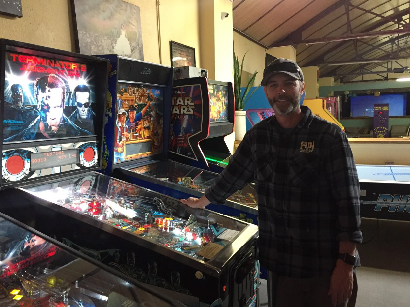 Man standing next to pinball machines in arcade, smiling, wearing a cap and plaid shirt.