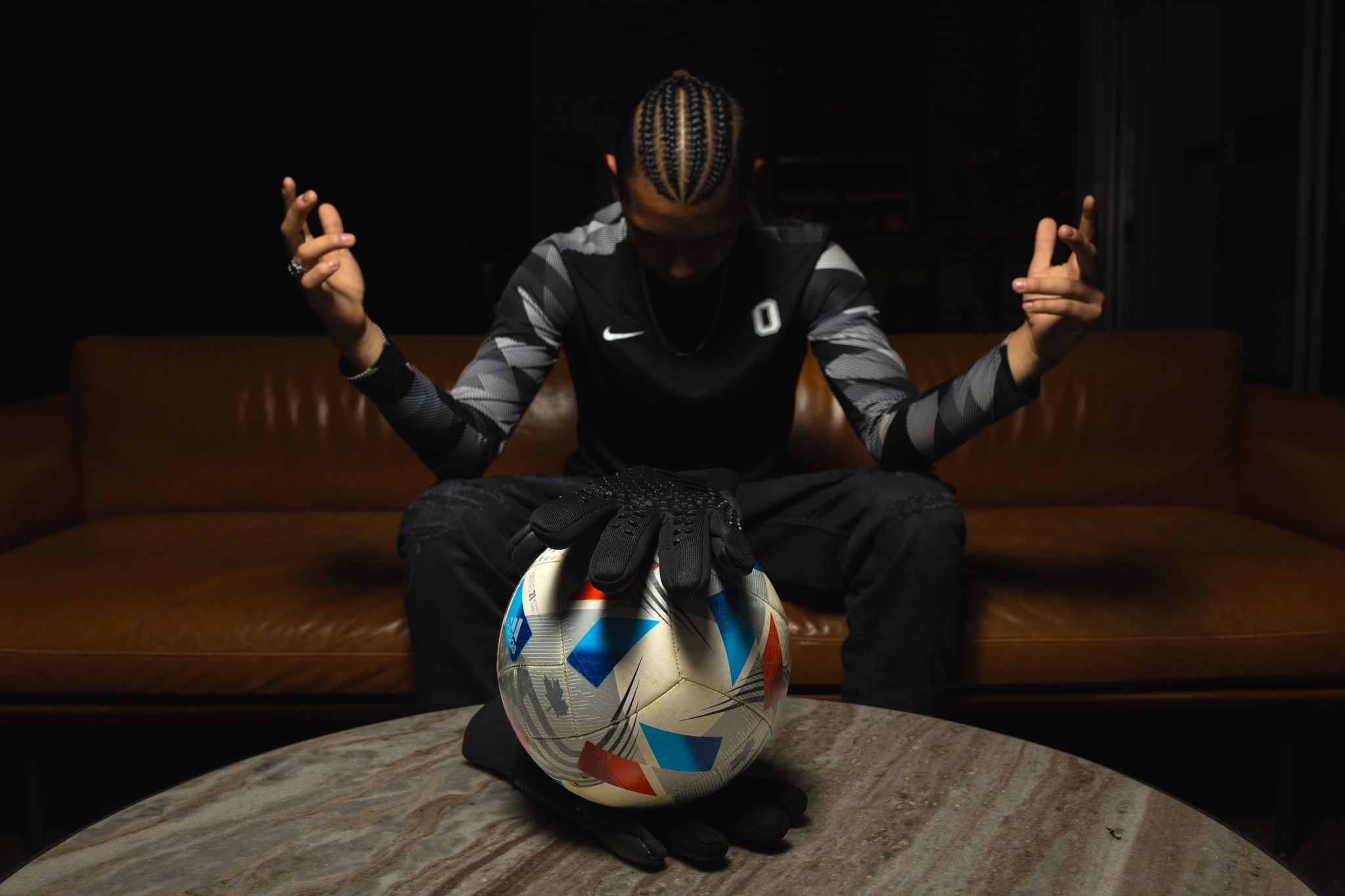 Person sitting on a couch with arms raised, wearing a sports jersey, with a colorful soccer ball on the table in front.