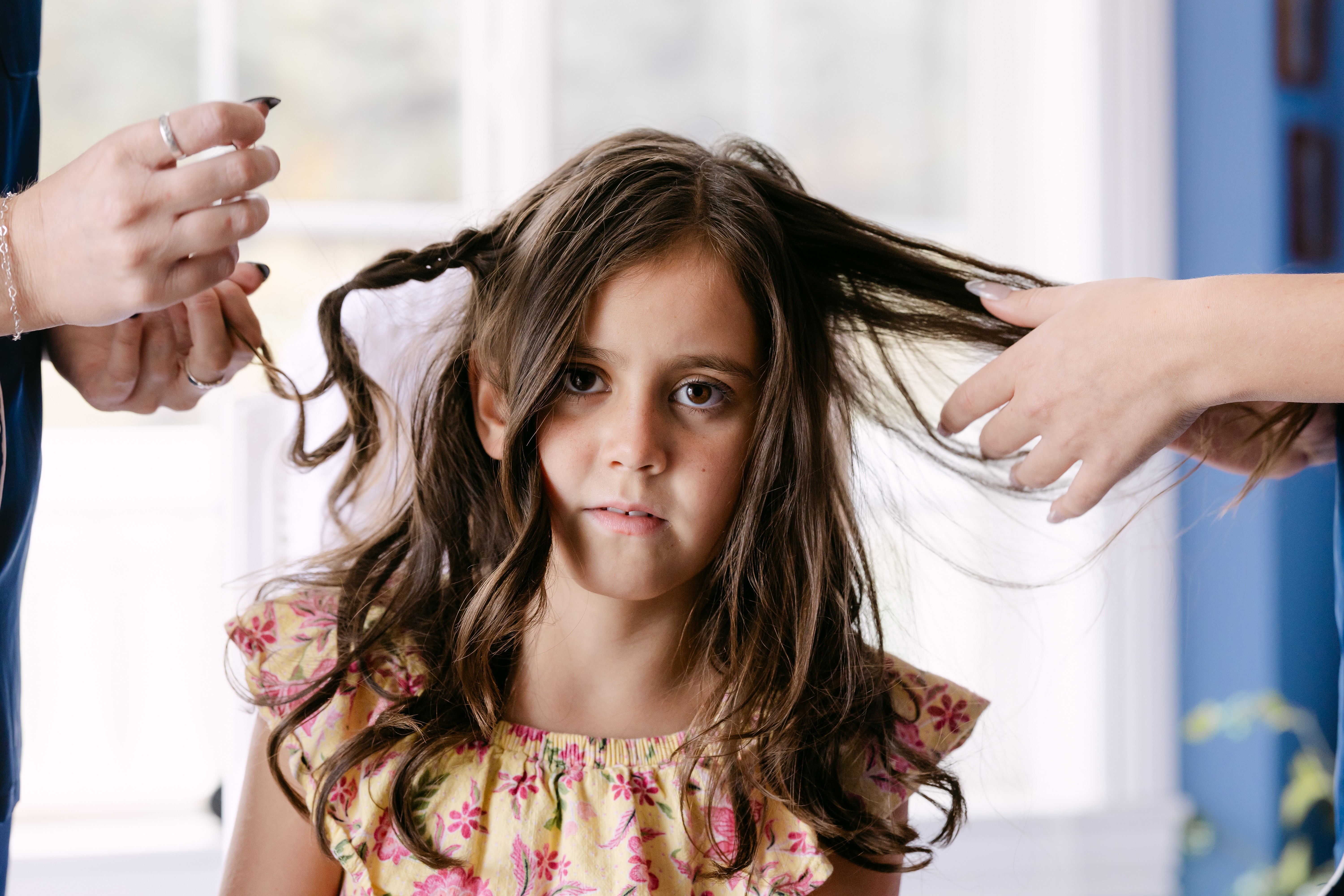 Young girl with long, wavy hair being styled by two people in a bright room with large windows.