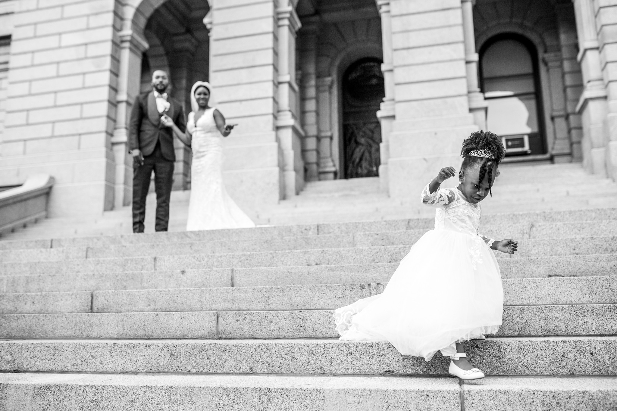 A bride in a white dress descends stairs, with a couple in formal attire in the background on the steps outside a building.