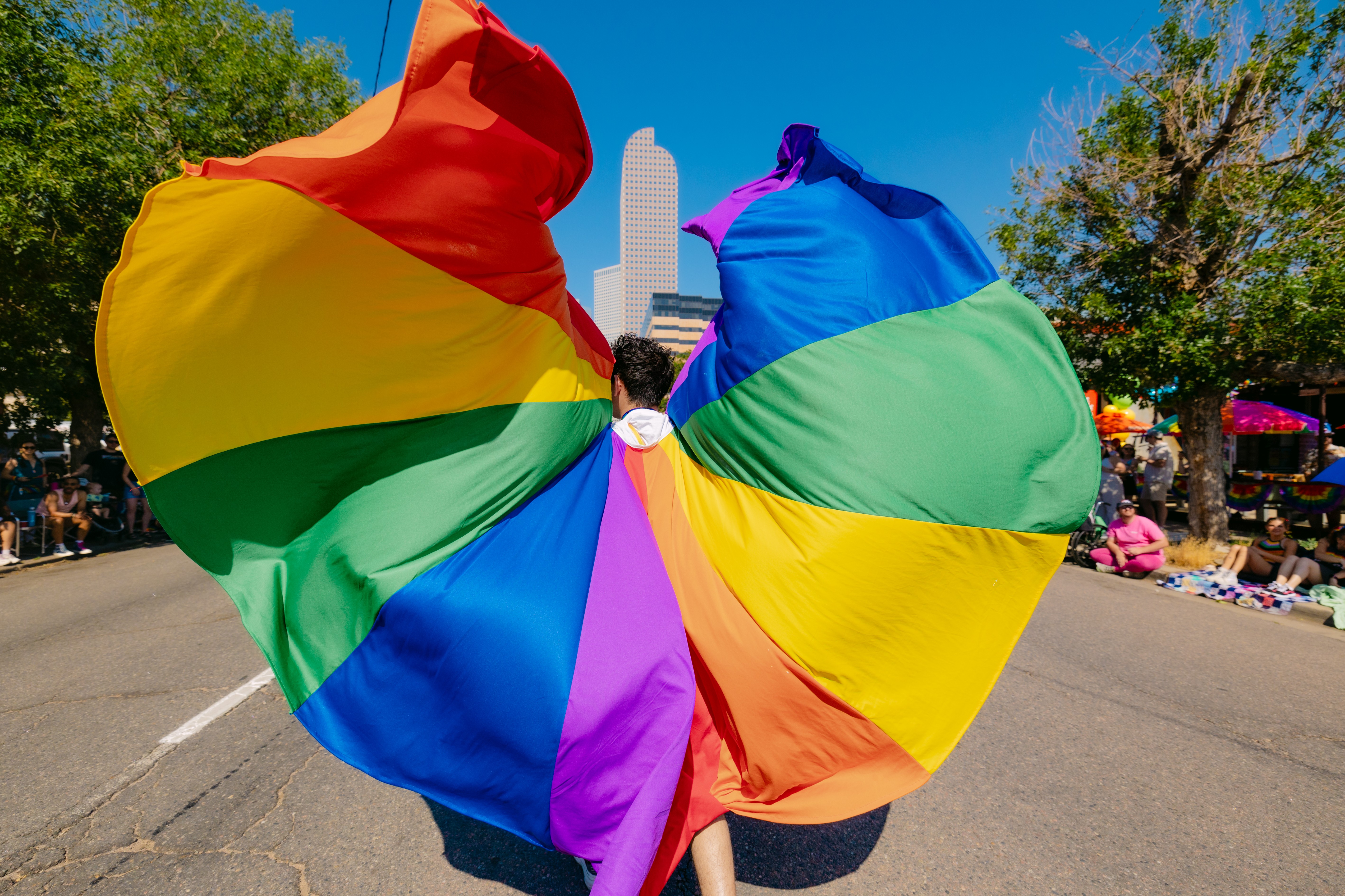 Person holding large rainbow flag at parade, with city skyline and trees in background.