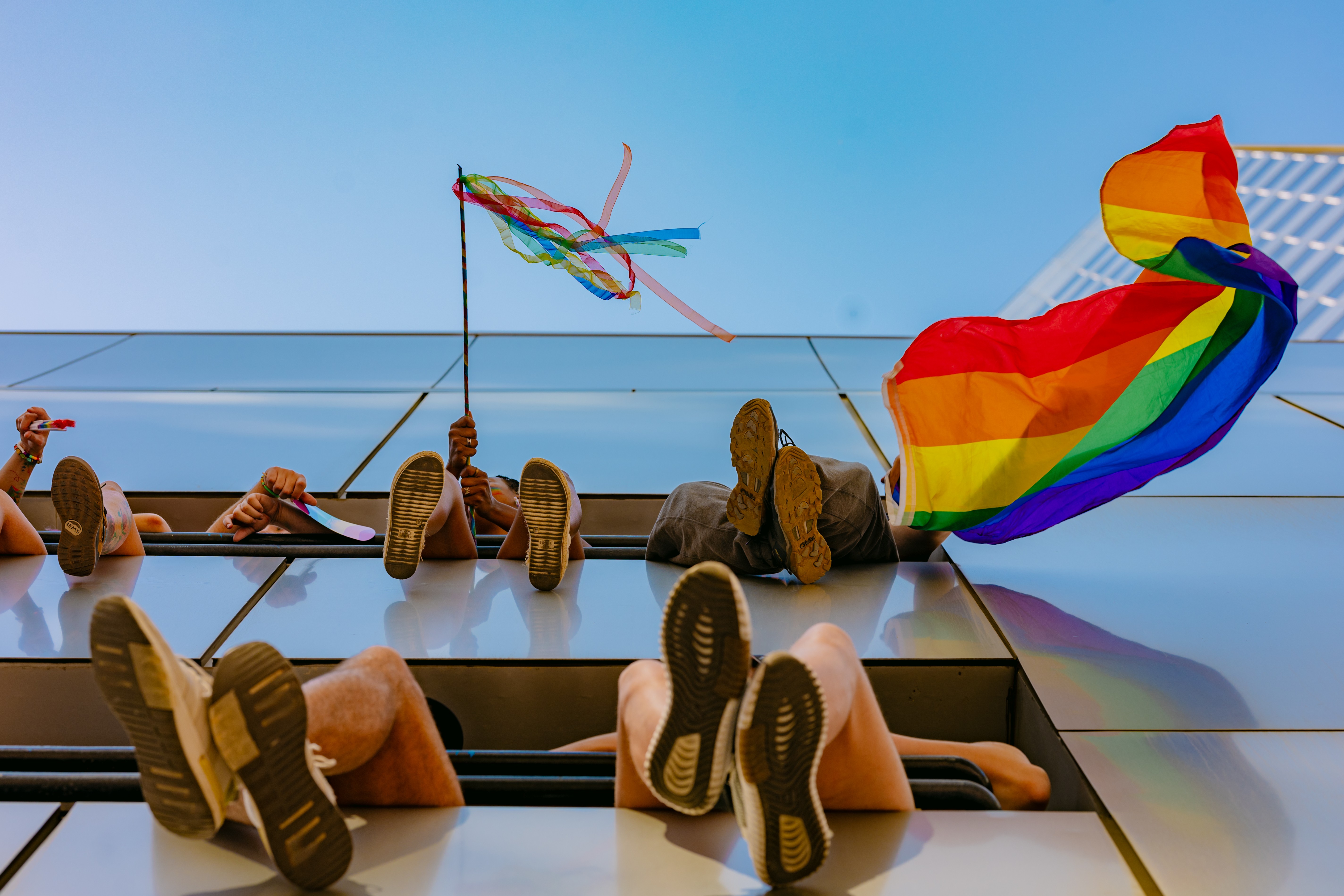 People lying on the ground with legs up, holding rainbow flags and a kite, against a blue sky.