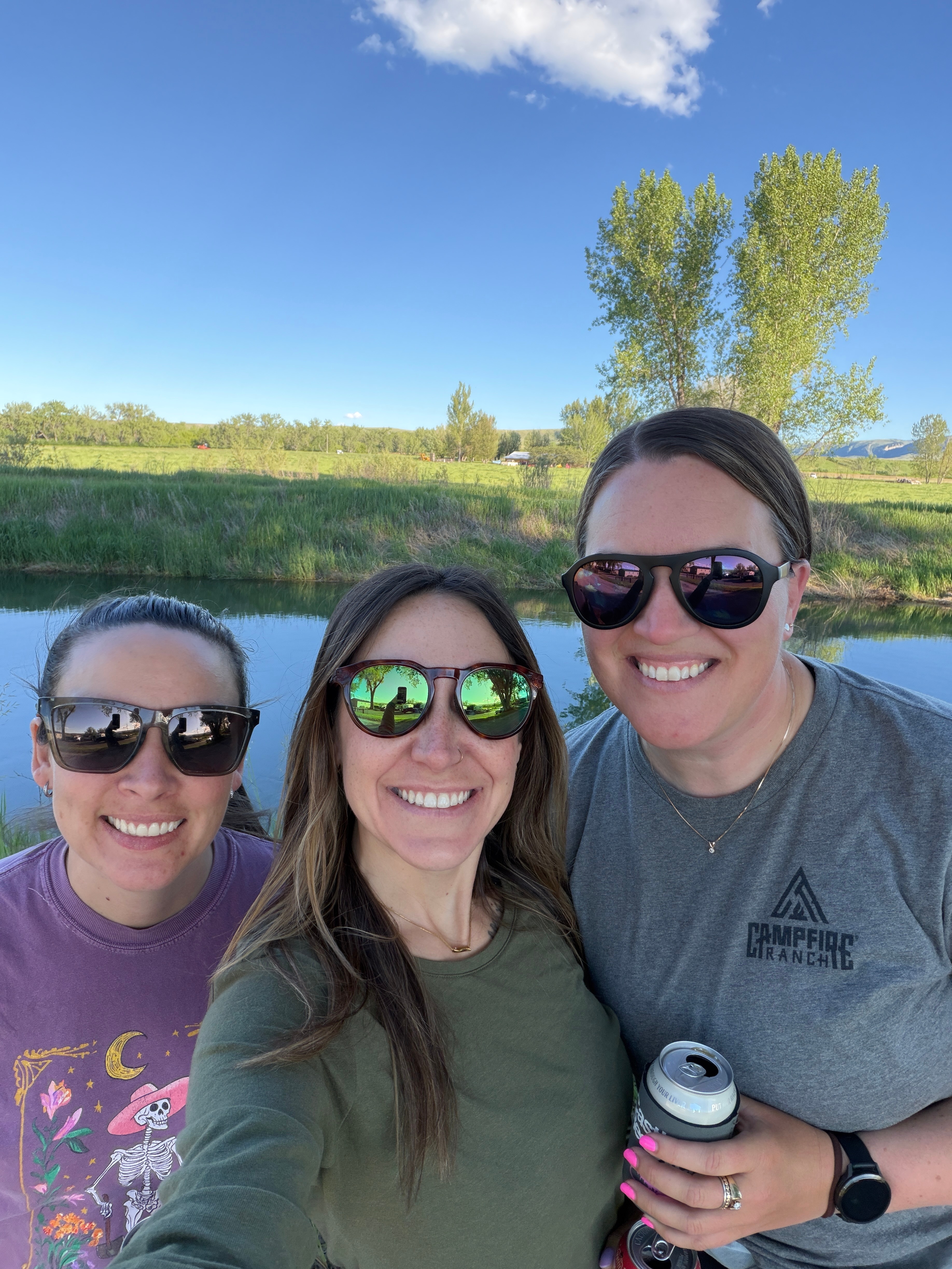 Three women outdoors near a river, smiling, wearing sunglasses, with trees and blue sky in background.