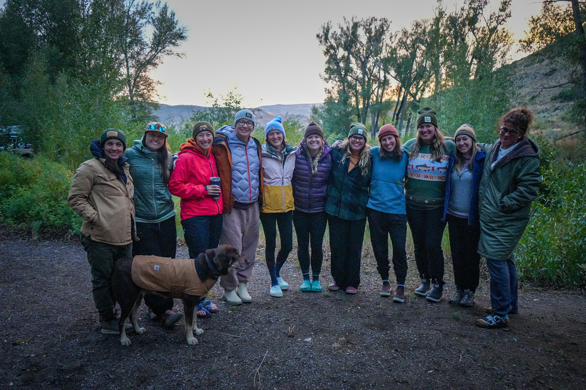 Group of ten people standing outdoors with trees and sky in background, some wearing jackets, one person with a dog.