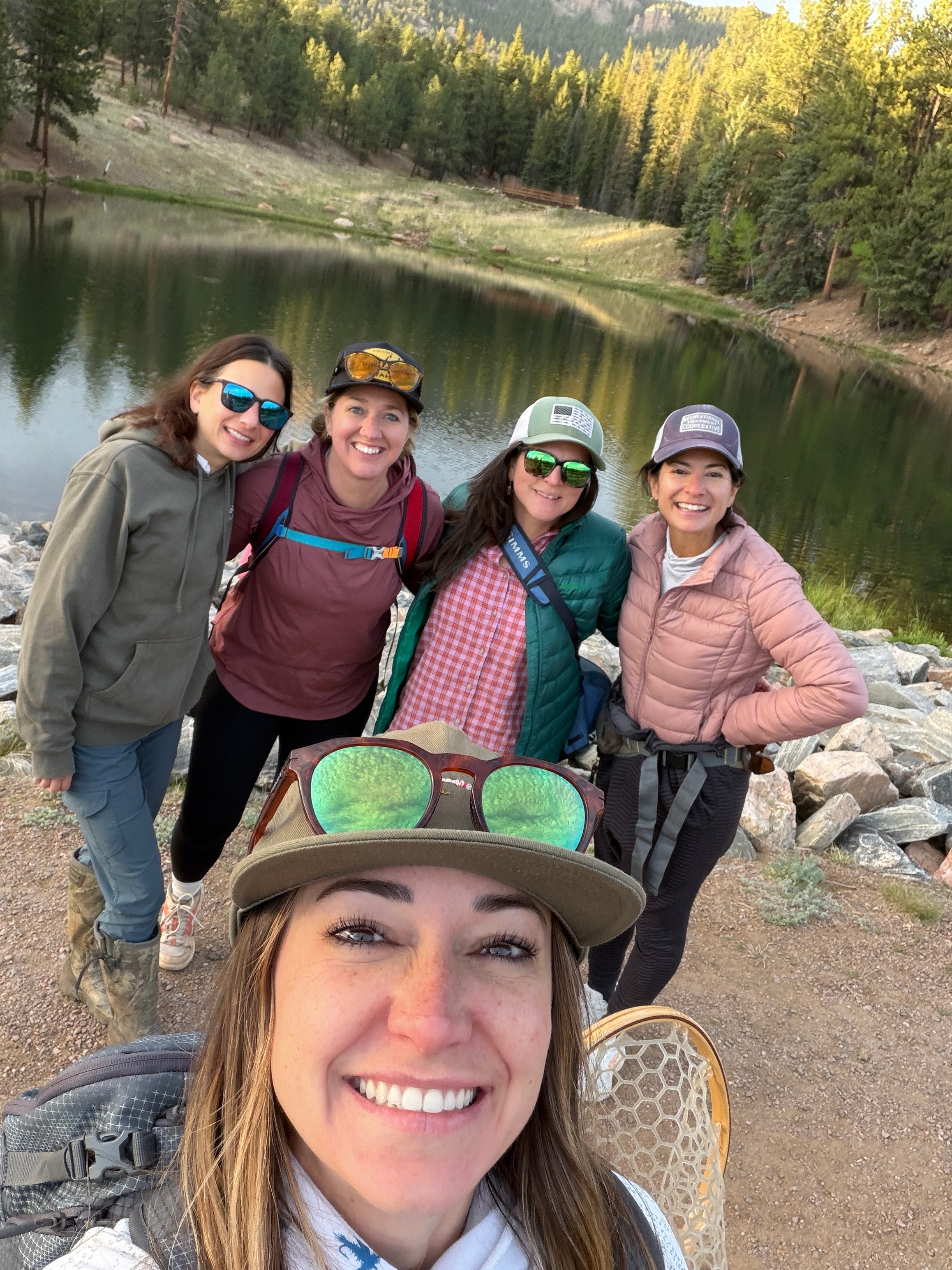 Four women outdoors near a lake, smiling, wearing sunglasses and casual clothing, with trees and water in the background.