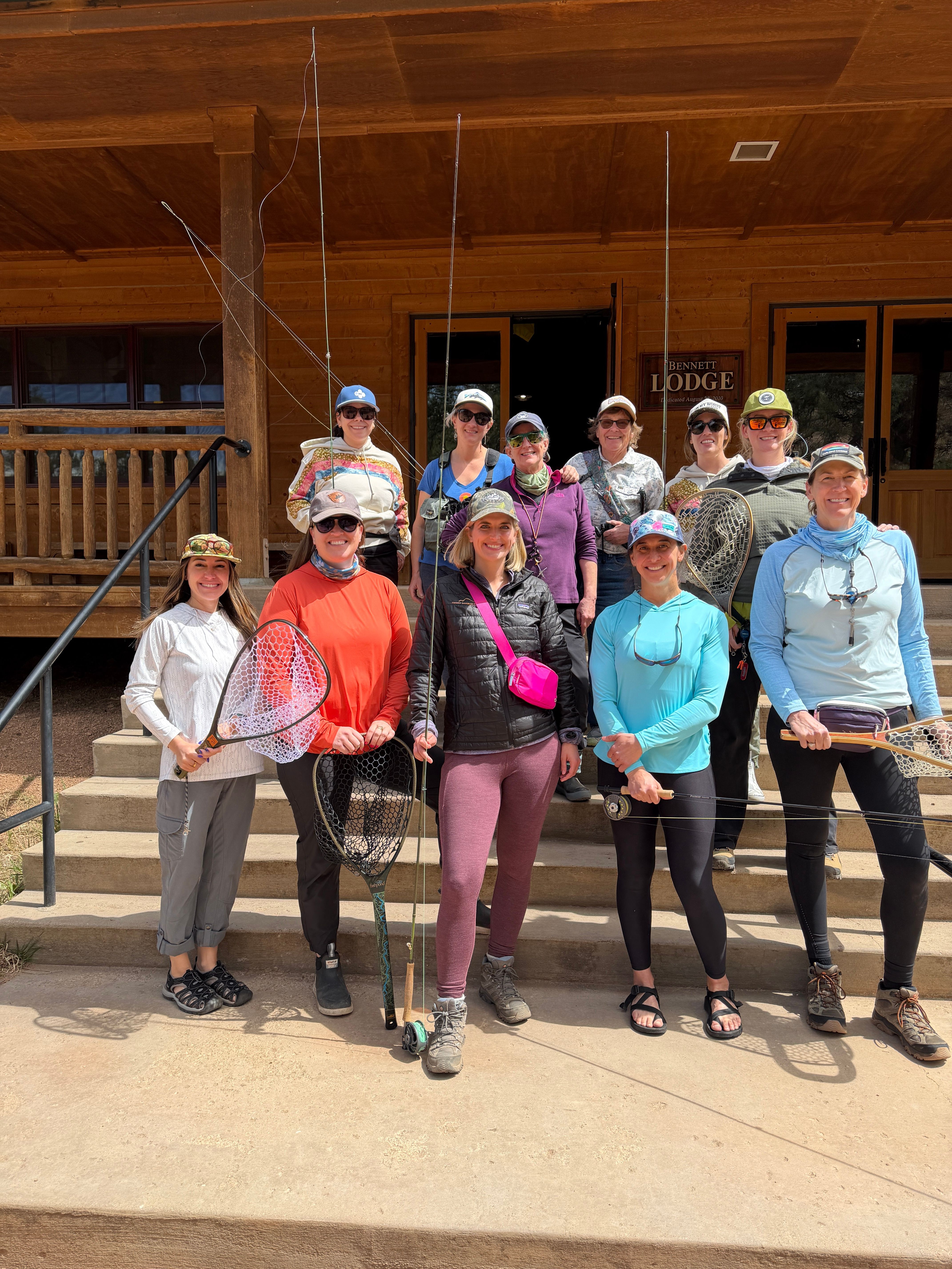 Group of people standing indoors, some holding fishing rods, posing for a photo, with wooden walls in background.