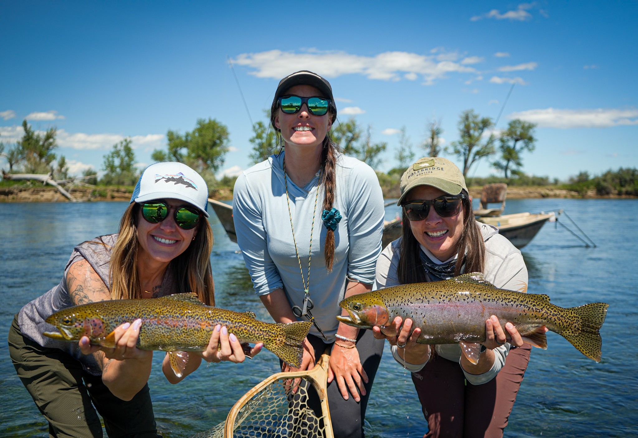 Three women outdoors near water holding two large fish, smiling, with trees and a boat in background.