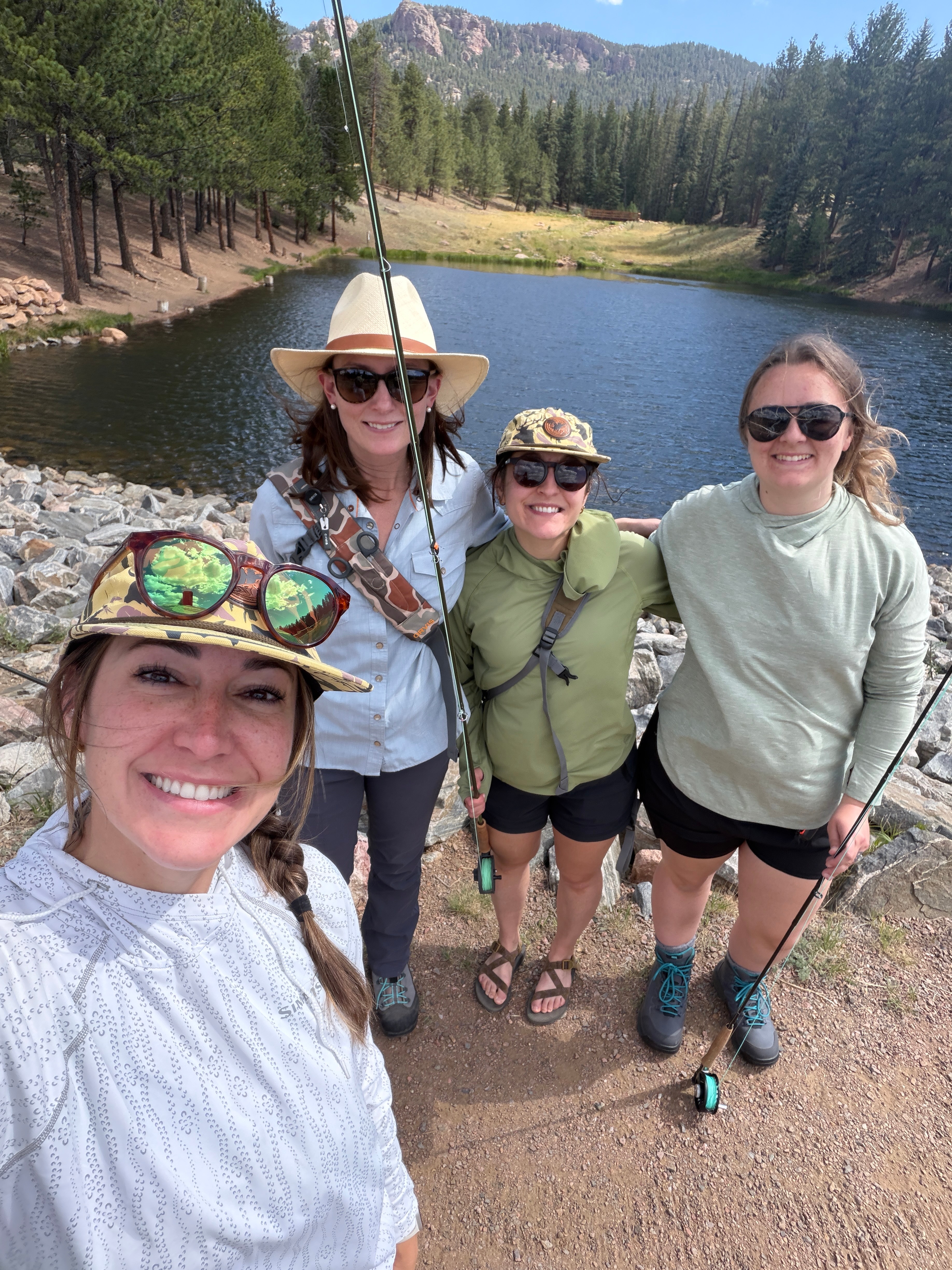 Four women outdoors near a lake with trees and mountains in the background, smiling and posing for a photo.