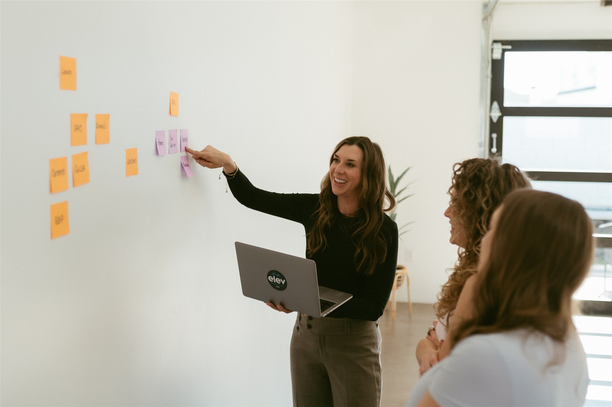 Woman presenting sticky notes on a whiteboard to three people in a bright room with a window.