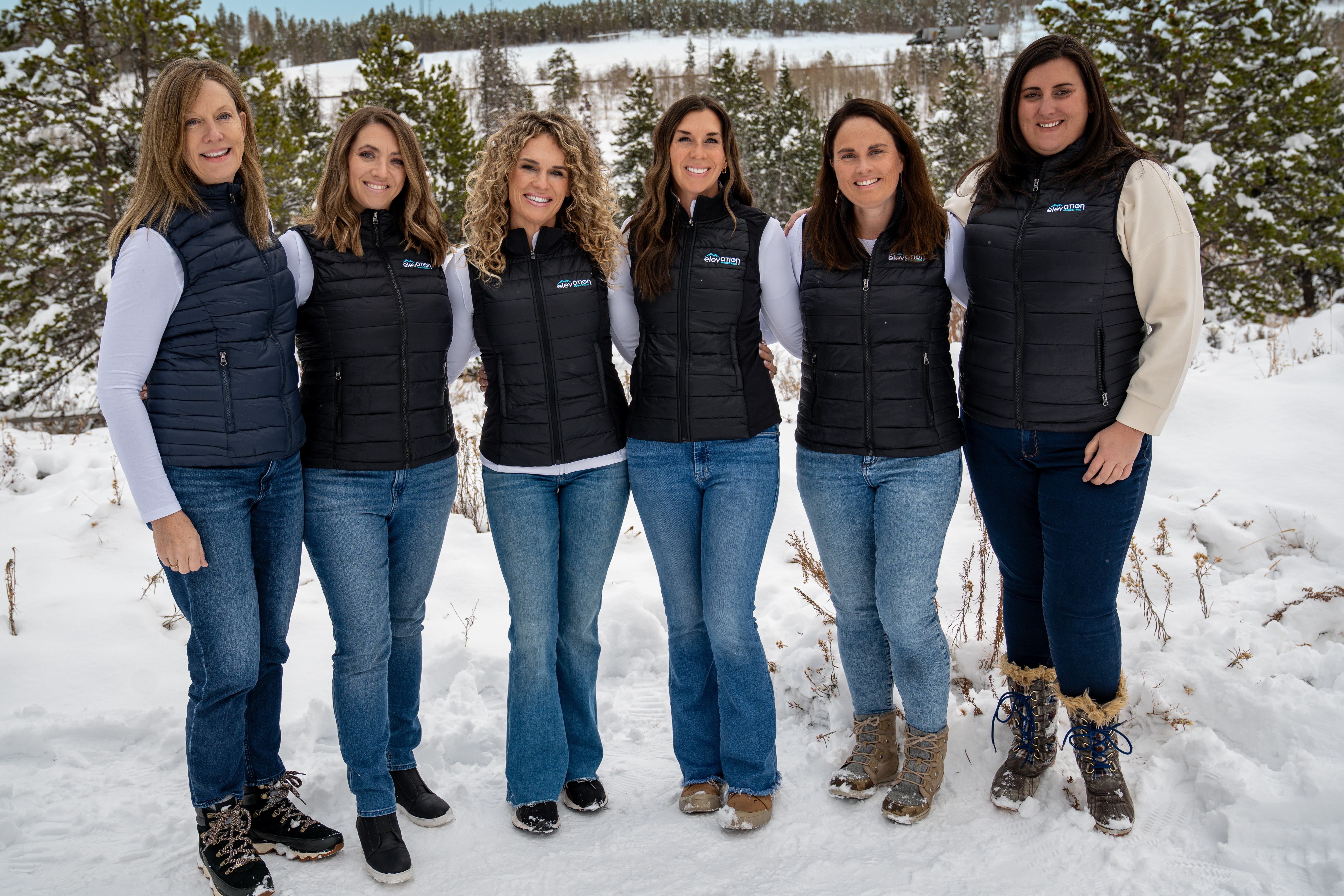 Six women stand outdoors in snow, wearing black vests and jeans, with trees and snow-covered landscape behind them.