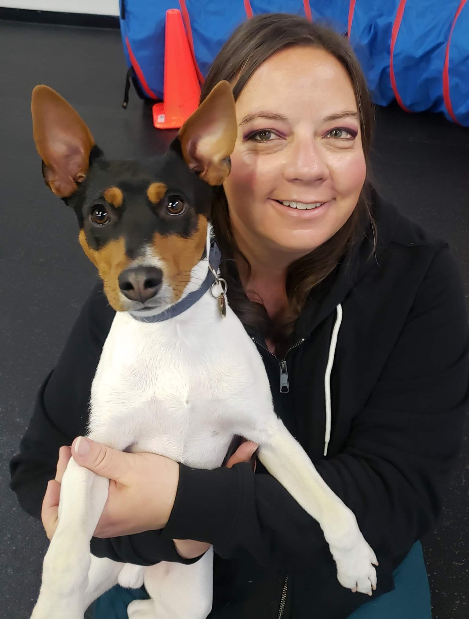 Woman holding a small dog with large ears, smiling, indoors. Background includes colorful objects. 20 words.