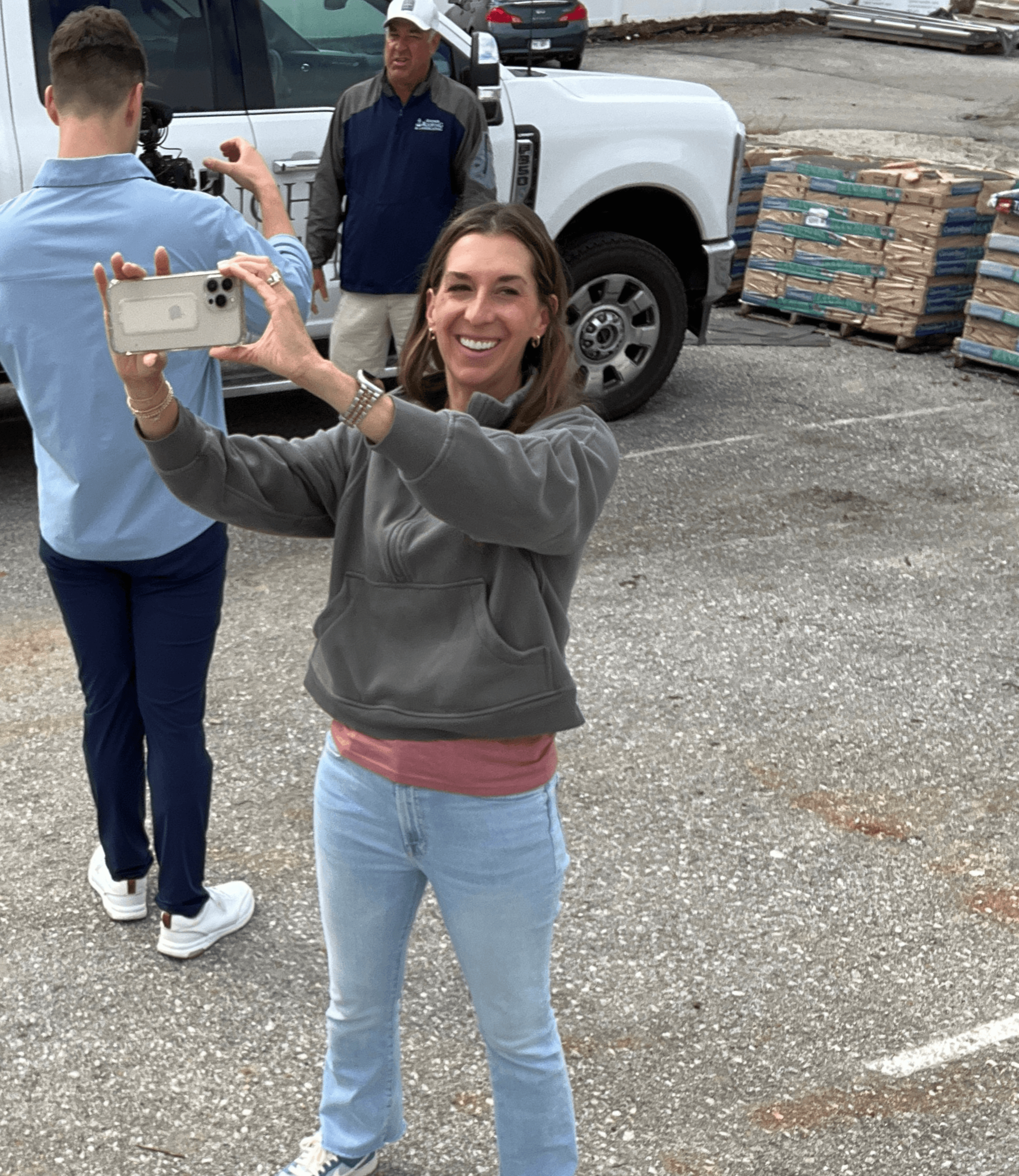 Woman smiling and taking a selfie outdoors with a smartphone, people and a white vehicle in the background.