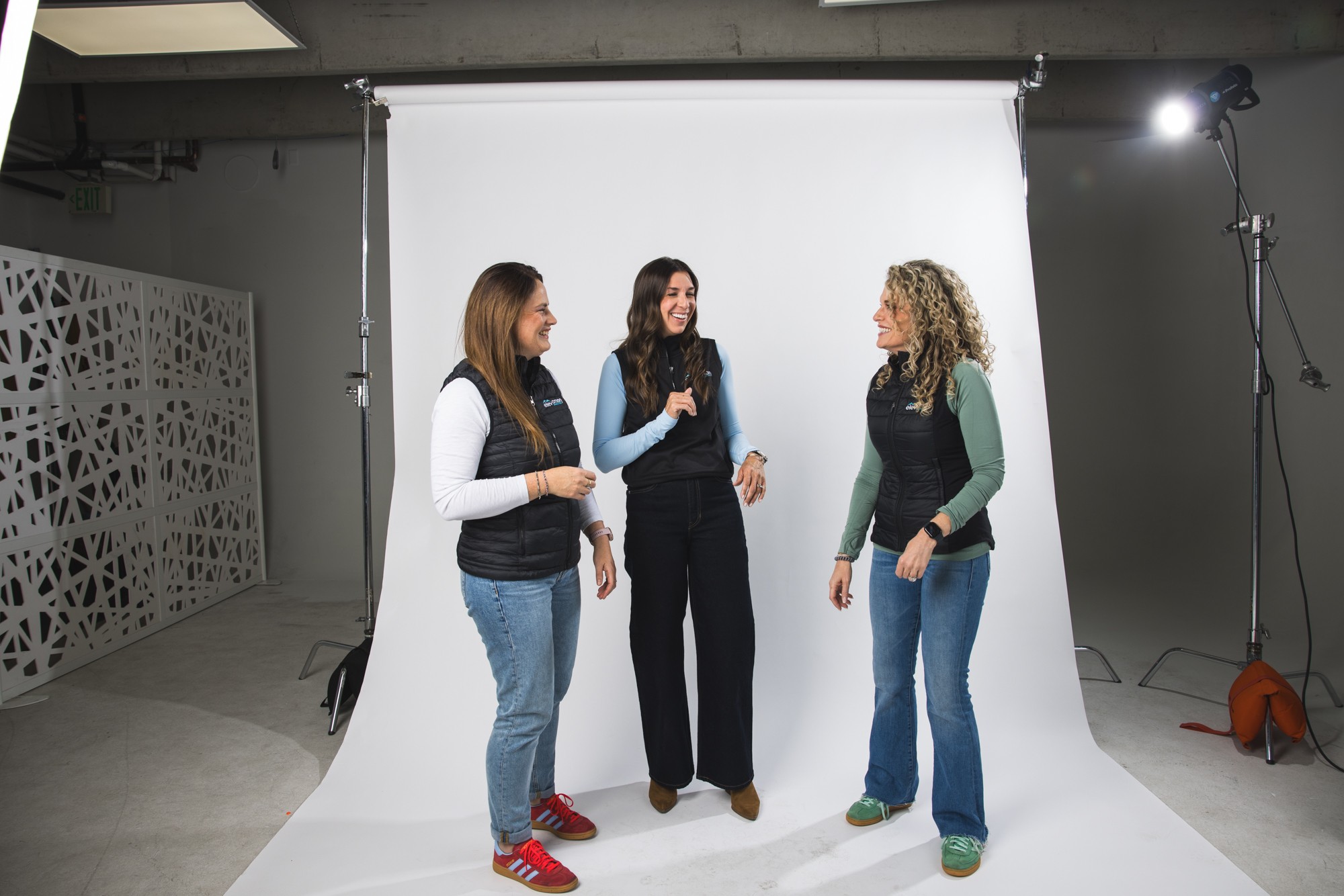 Three women standing and talking in a photography studio with a white backdrop, lighting equipment, and a patterned wall.