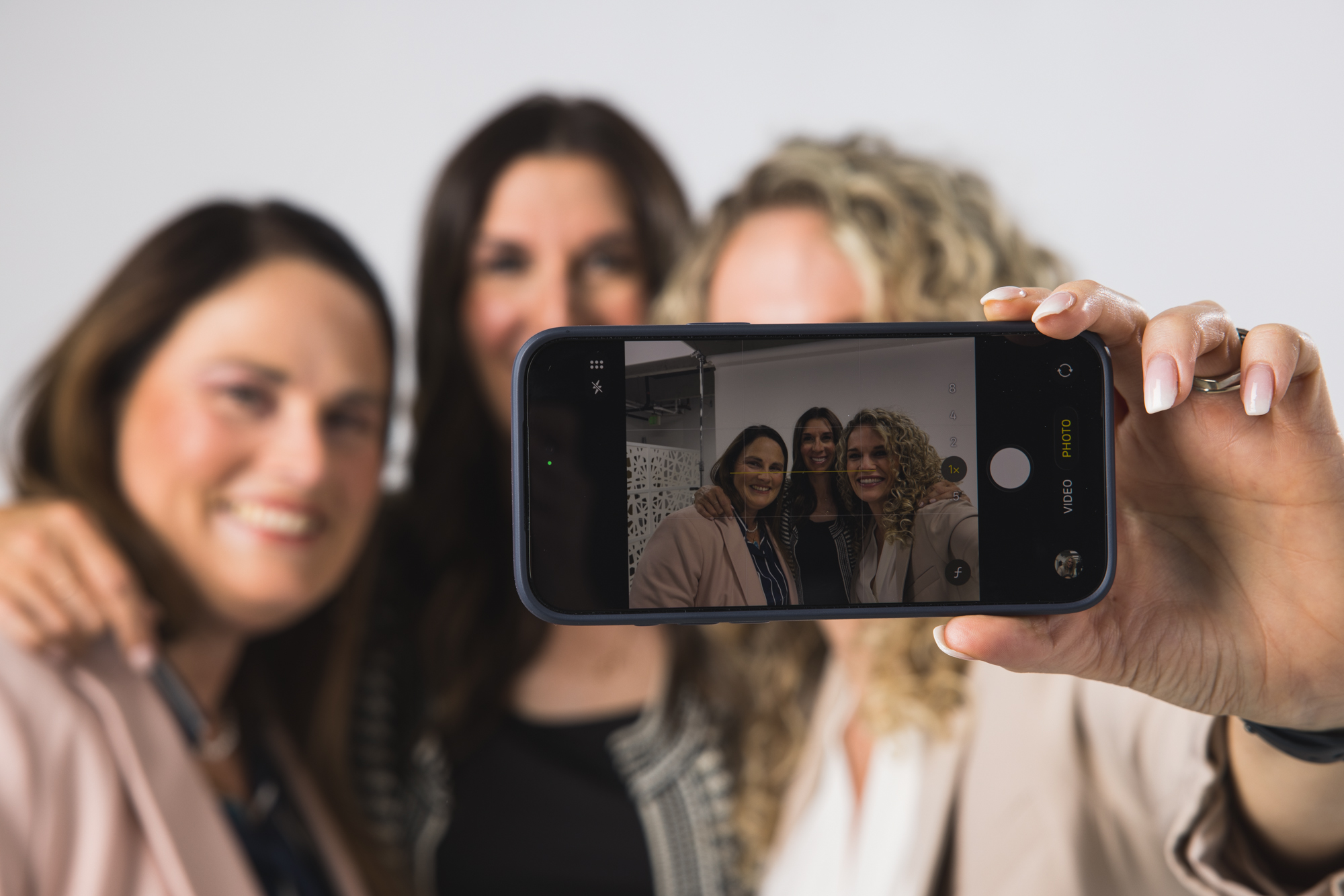 Person taking a selfie of three women smiling, with the phone visible in the foreground.