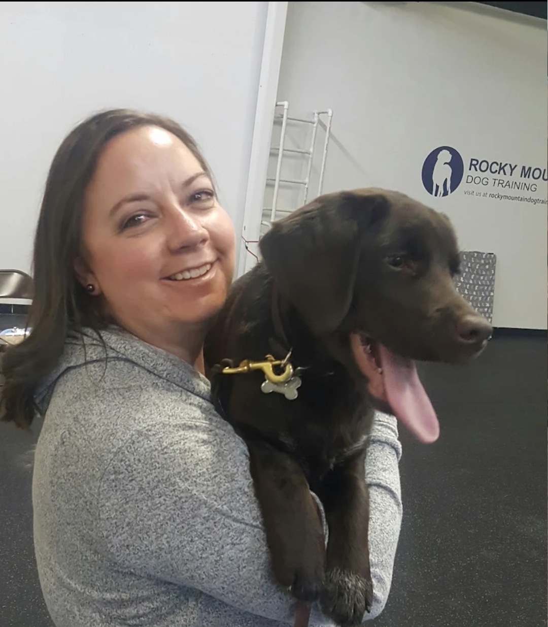 Woman smiling and holding a black puppy with its tongue out inside a training facility.