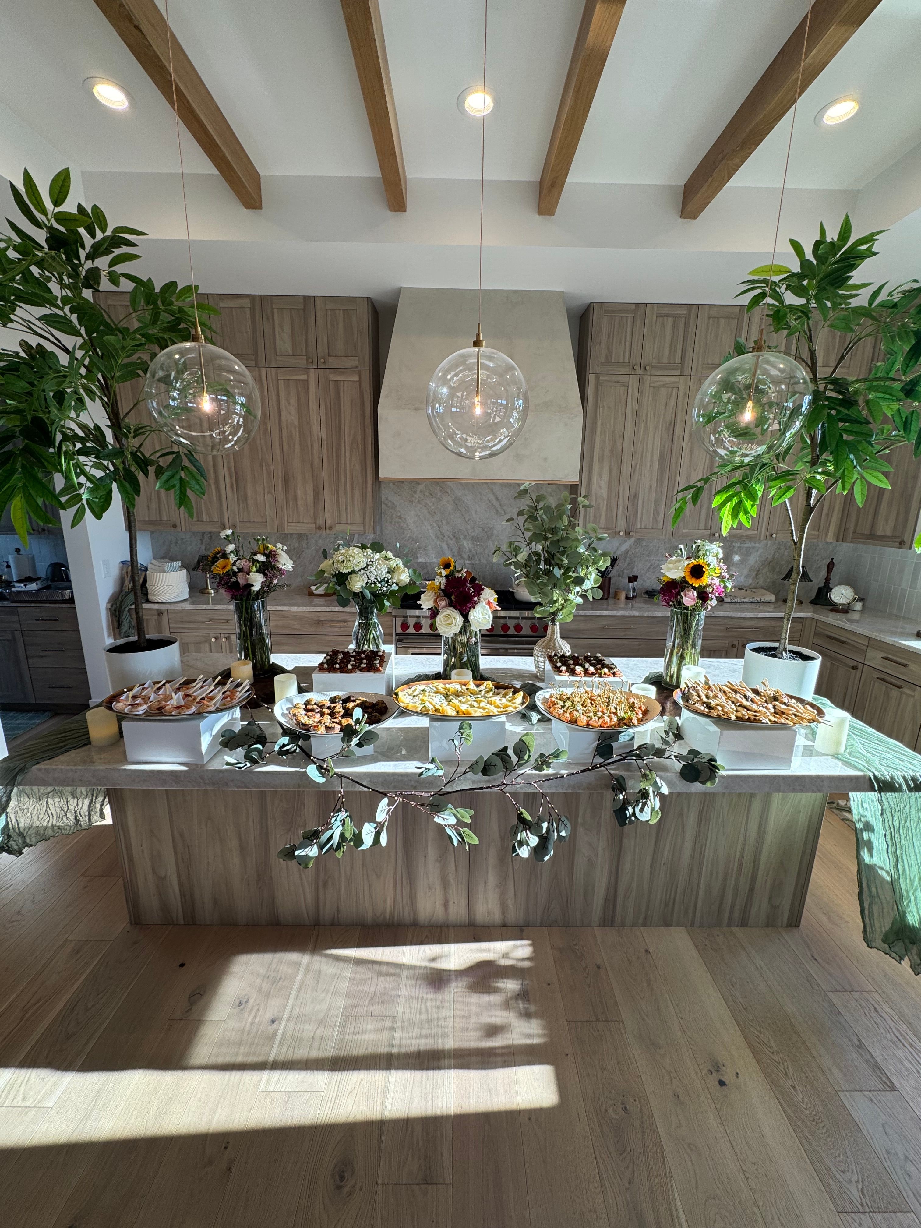 Table with floral arrangements, candles, and food, set in a bright room with wooden floors and shelves, sunlight streaming in.
