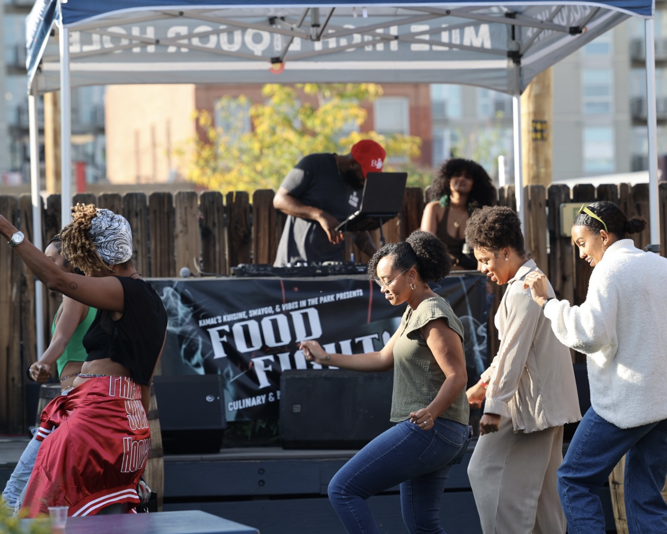 Group of people dancing outdoors under a canopy, with a food truck in the background.