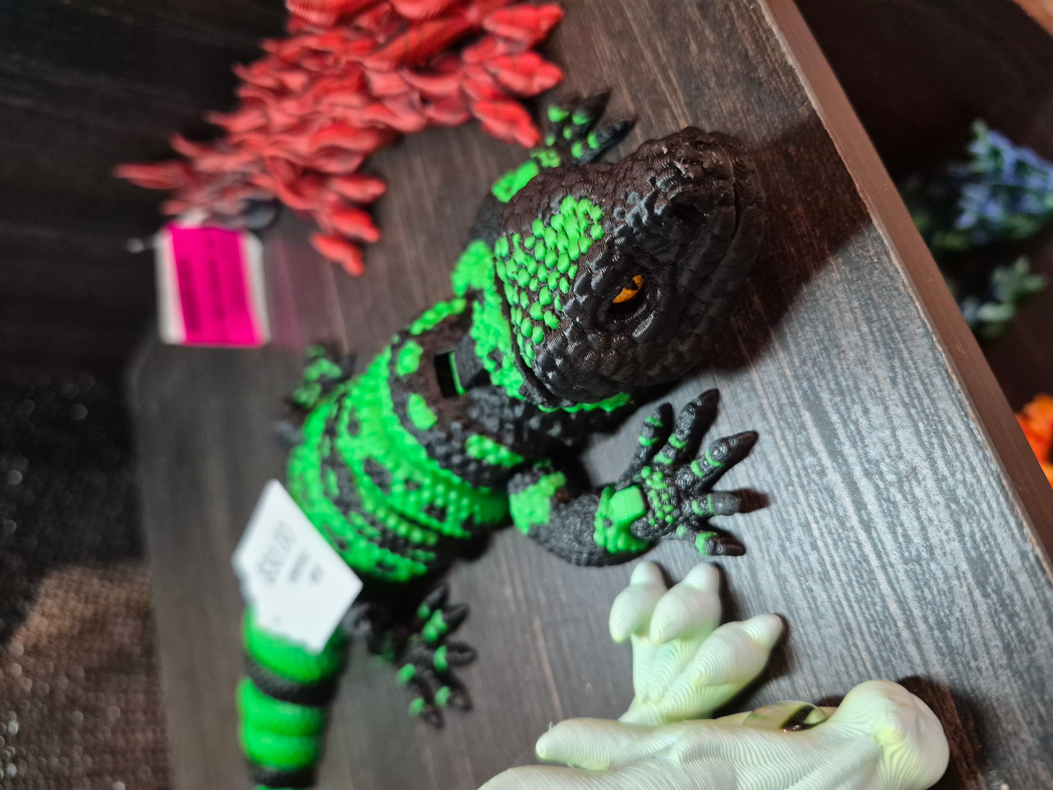 Green and black patterned lizard on wooden surface with white object nearby, pink and red items in background.