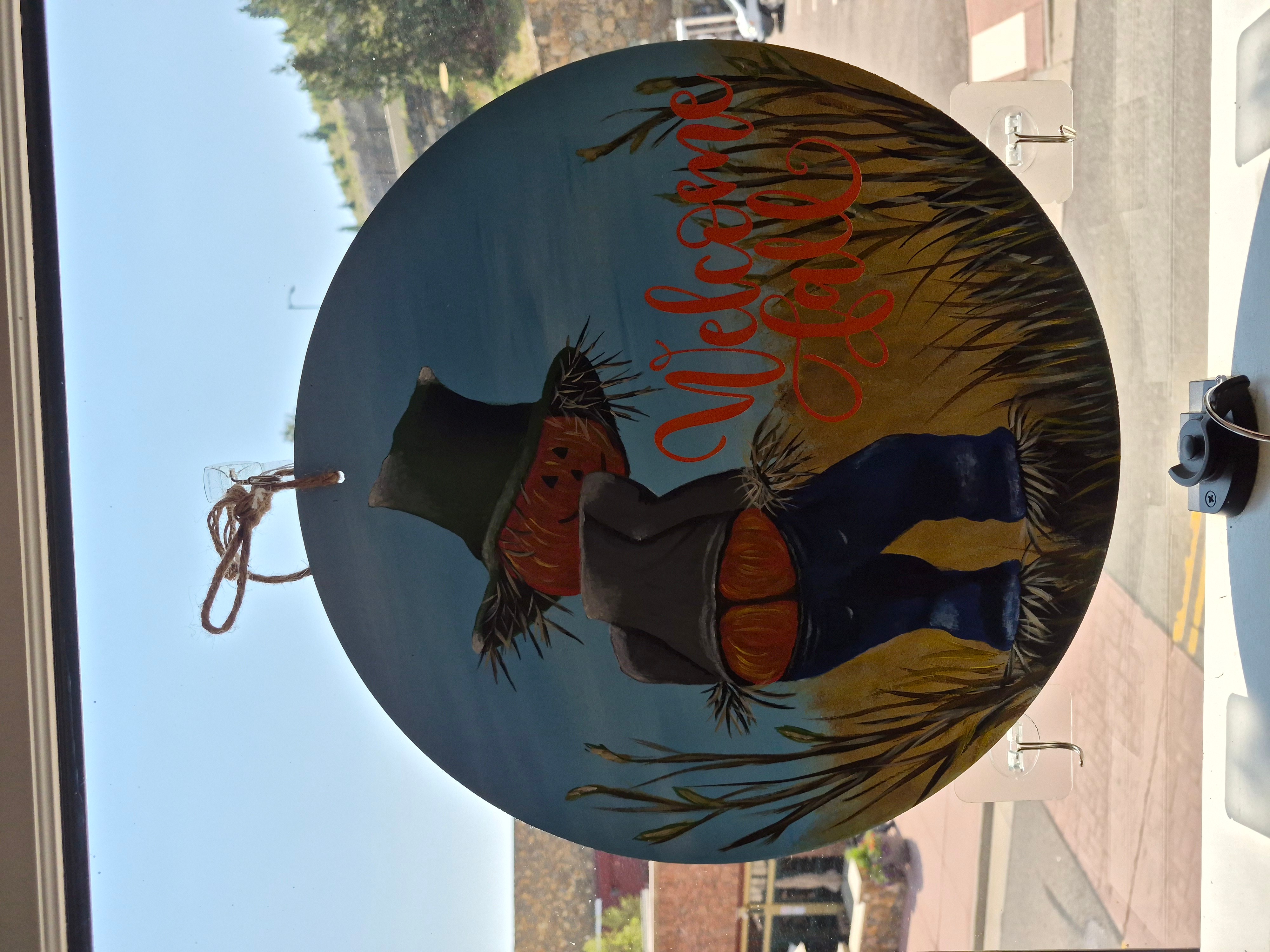 A circular sign with a scarecrow figure and red text, outdoors with buildings and sky in background.