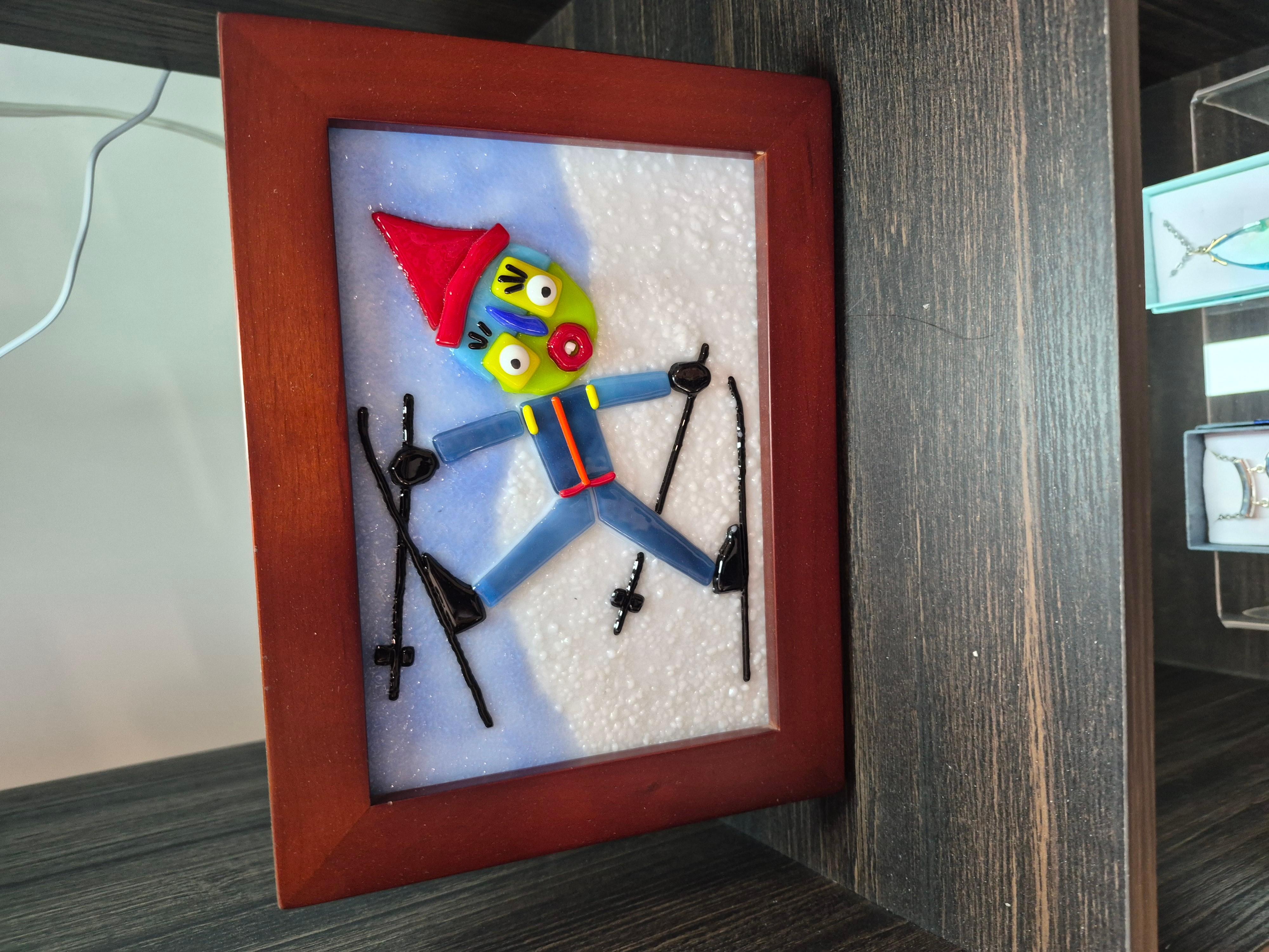 Colorful clown figure with a red hat and striped outfit, hanging inside a wooden frame on a wall.