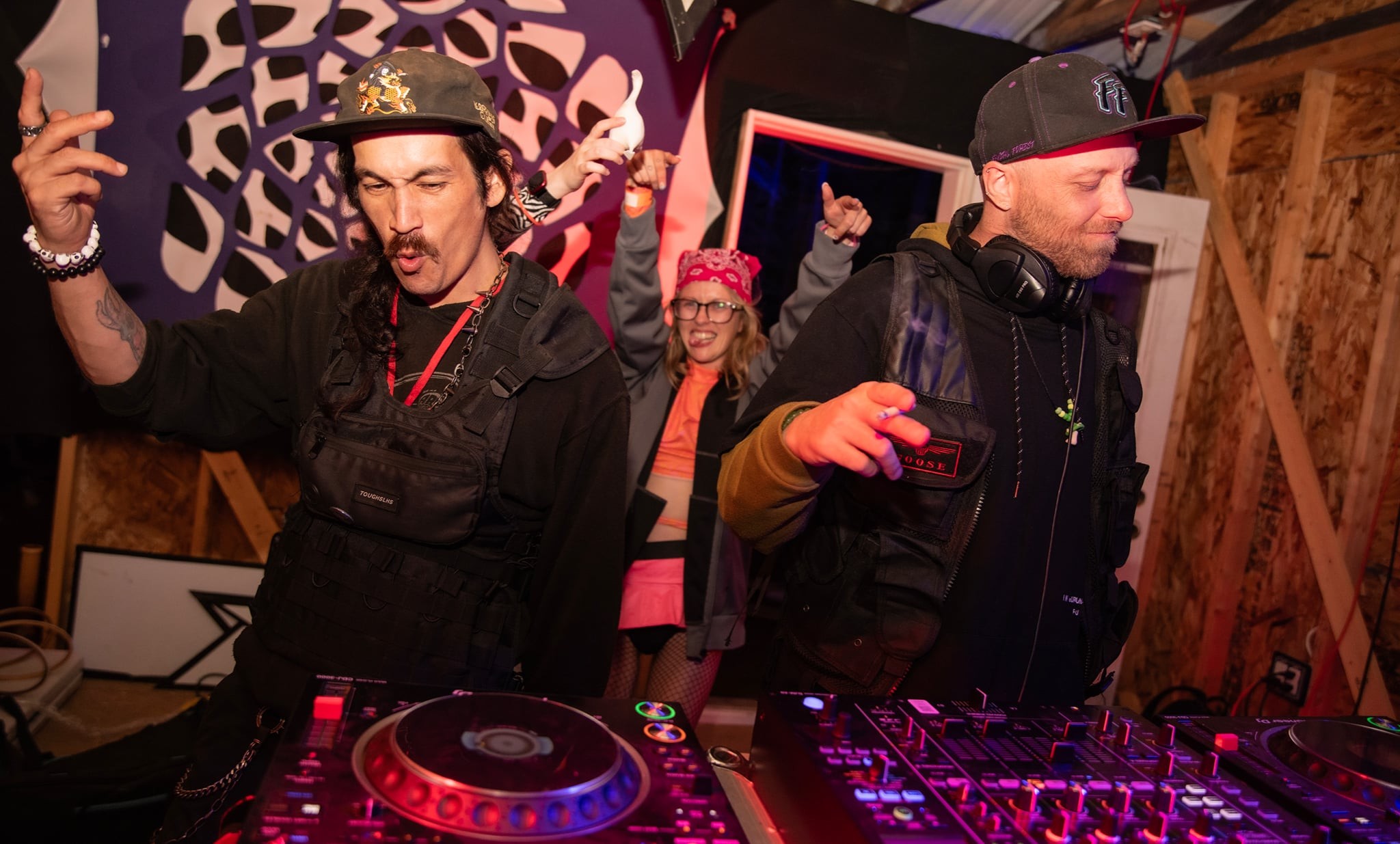 Two DJs wearing hats and headphones perform at a party with a woman in the background, wooden walls, and DJ equipment.