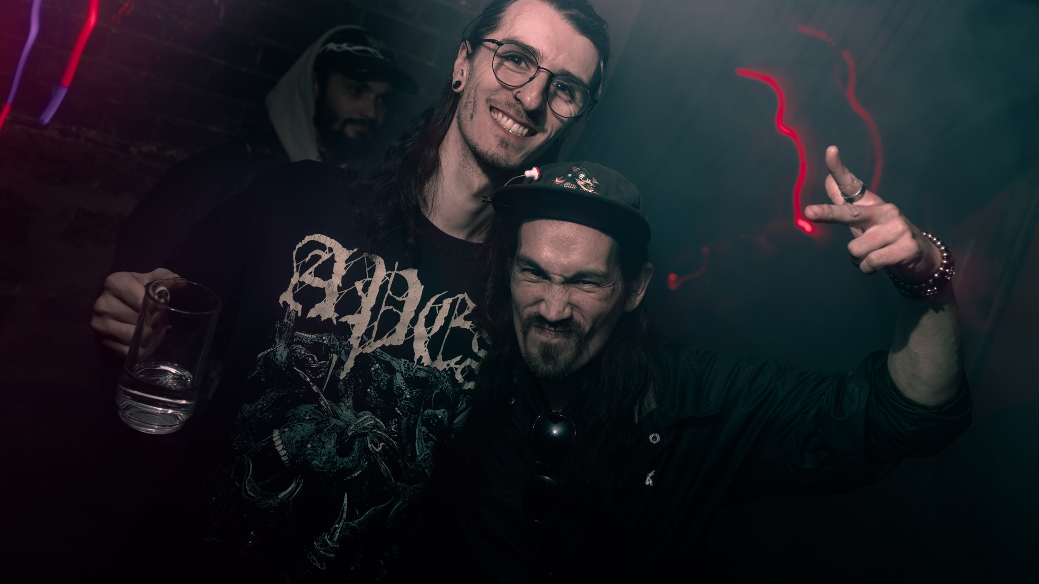 Two men smiling and making gestures in a dark, colorful environment, one holding a drink.
