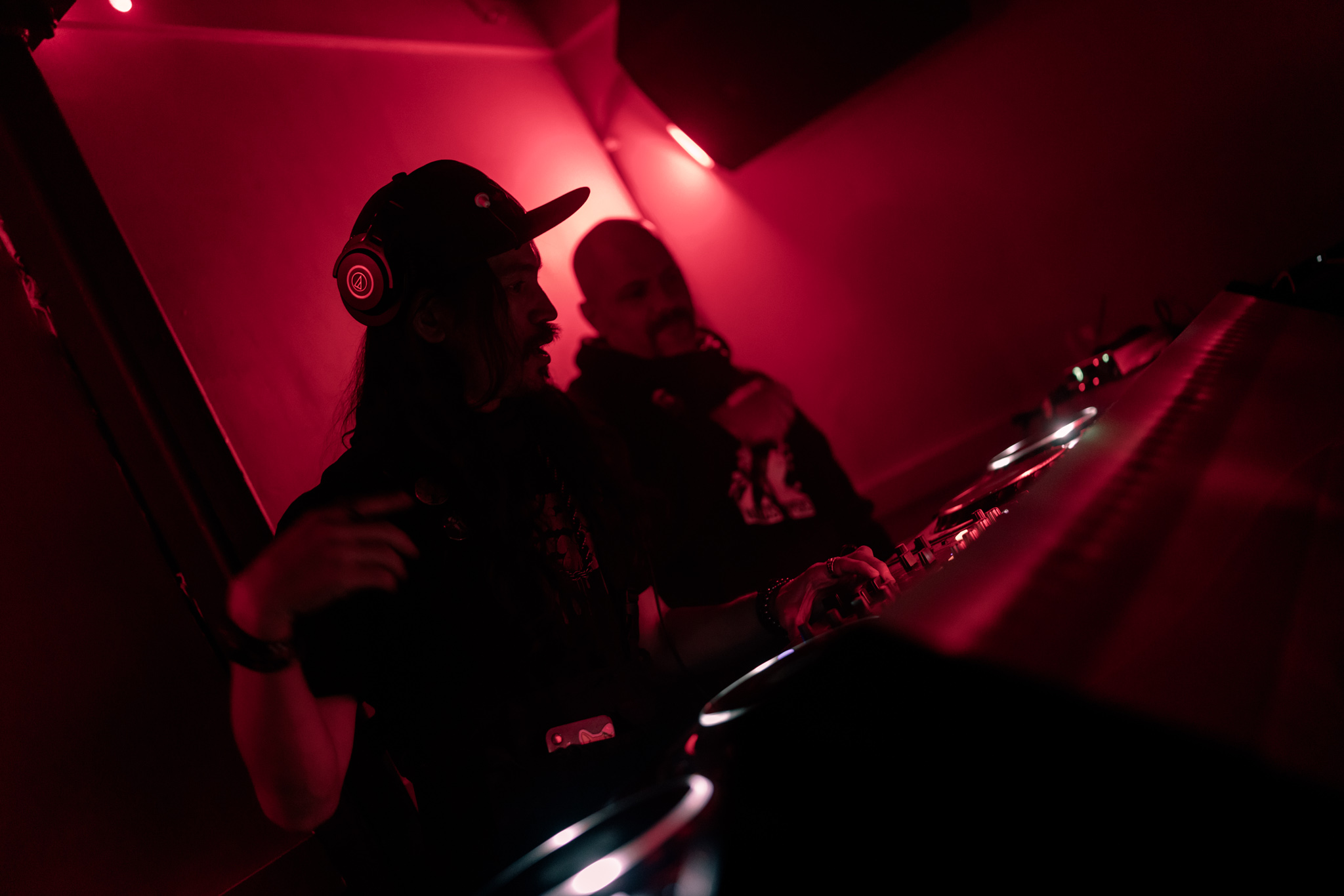 Two people in a dimly lit room with pink lighting, one wearing a cap, the other with a bun hairstyle, near a bar or DJ setup.