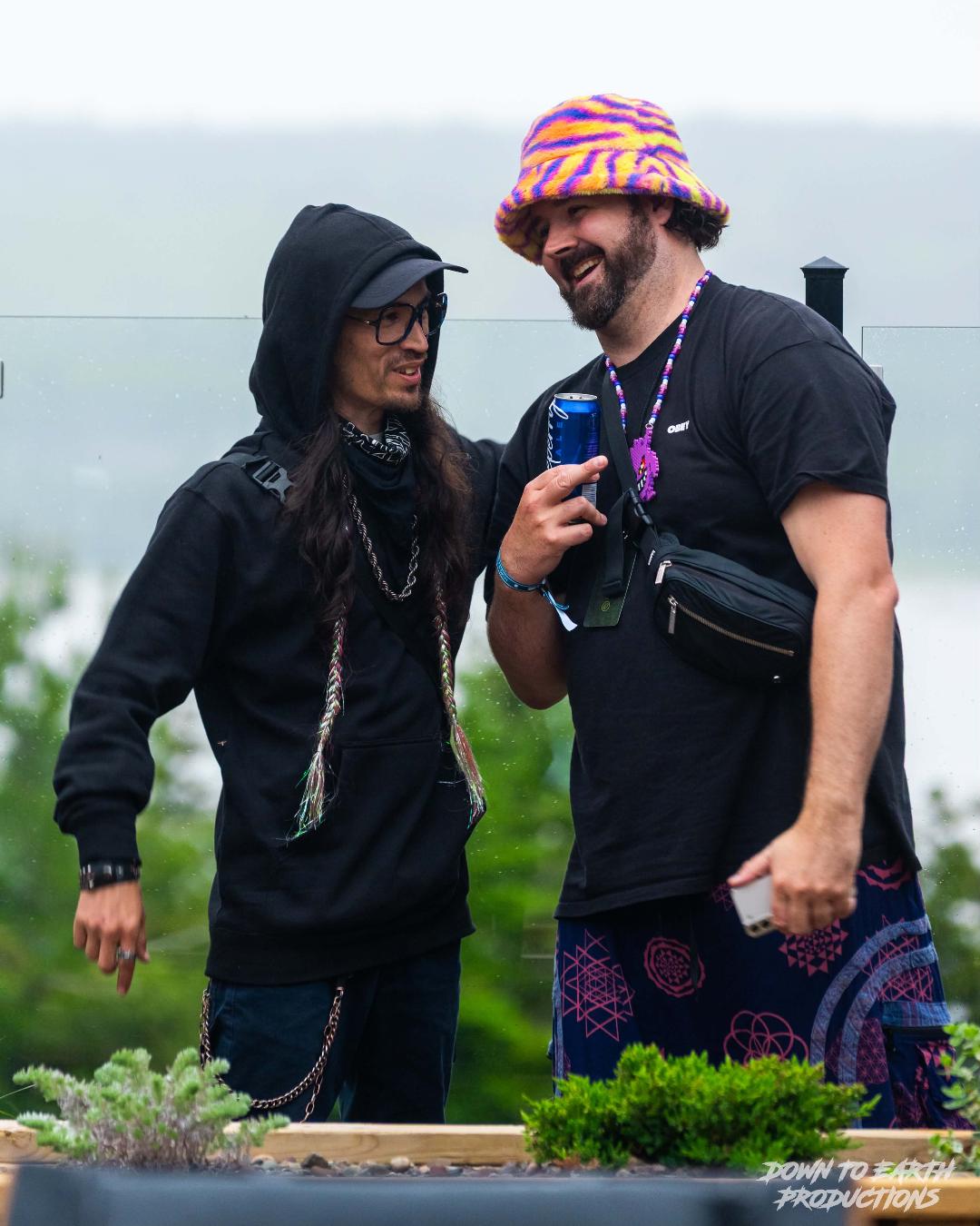 Two people outdoors, one with a hoodie and glasses, the other with a colorful hat, smiling and talking, greenery in background.