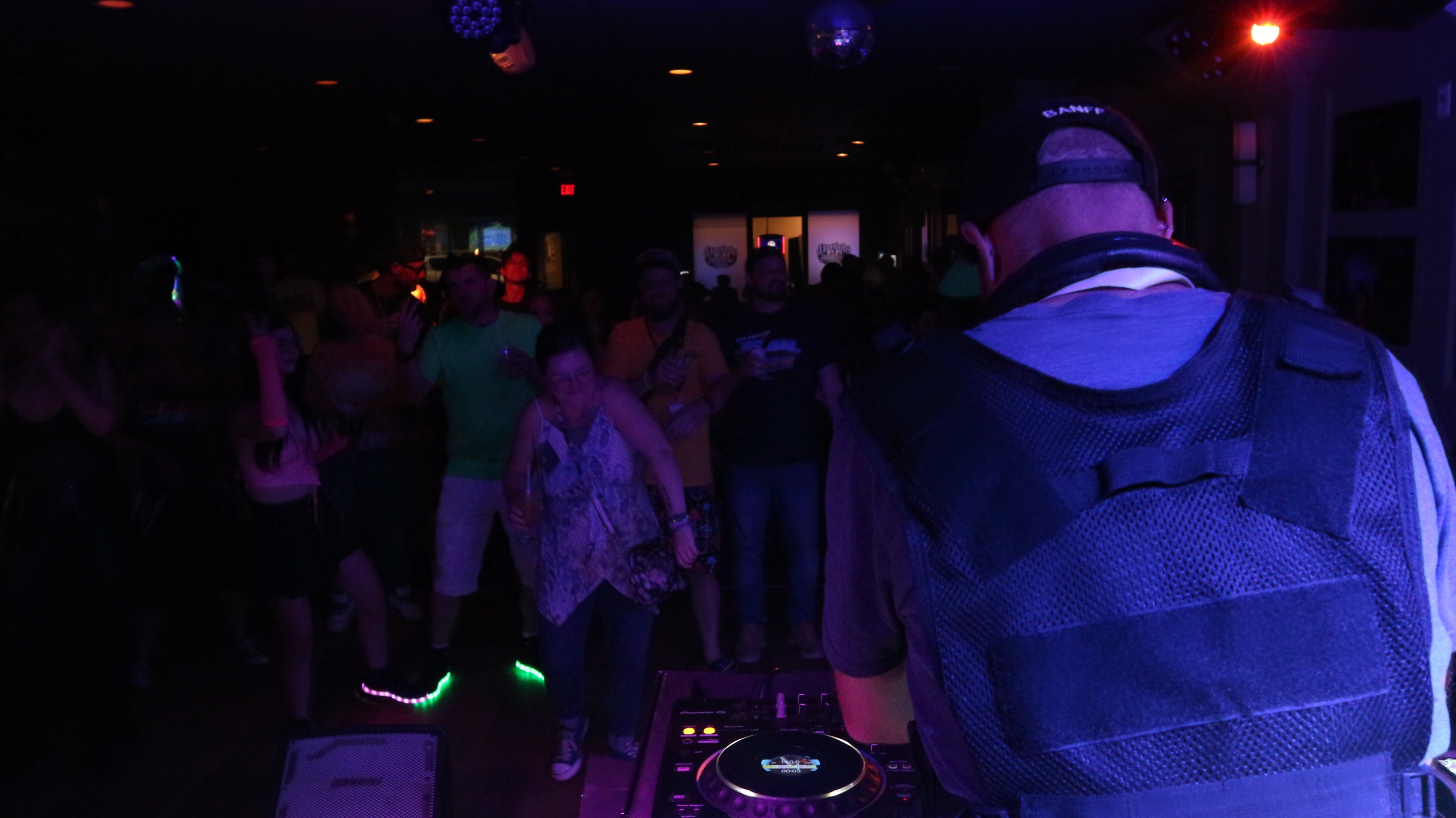 DJ in a dark club with people dancing and colorful lights, facing away from the camera.