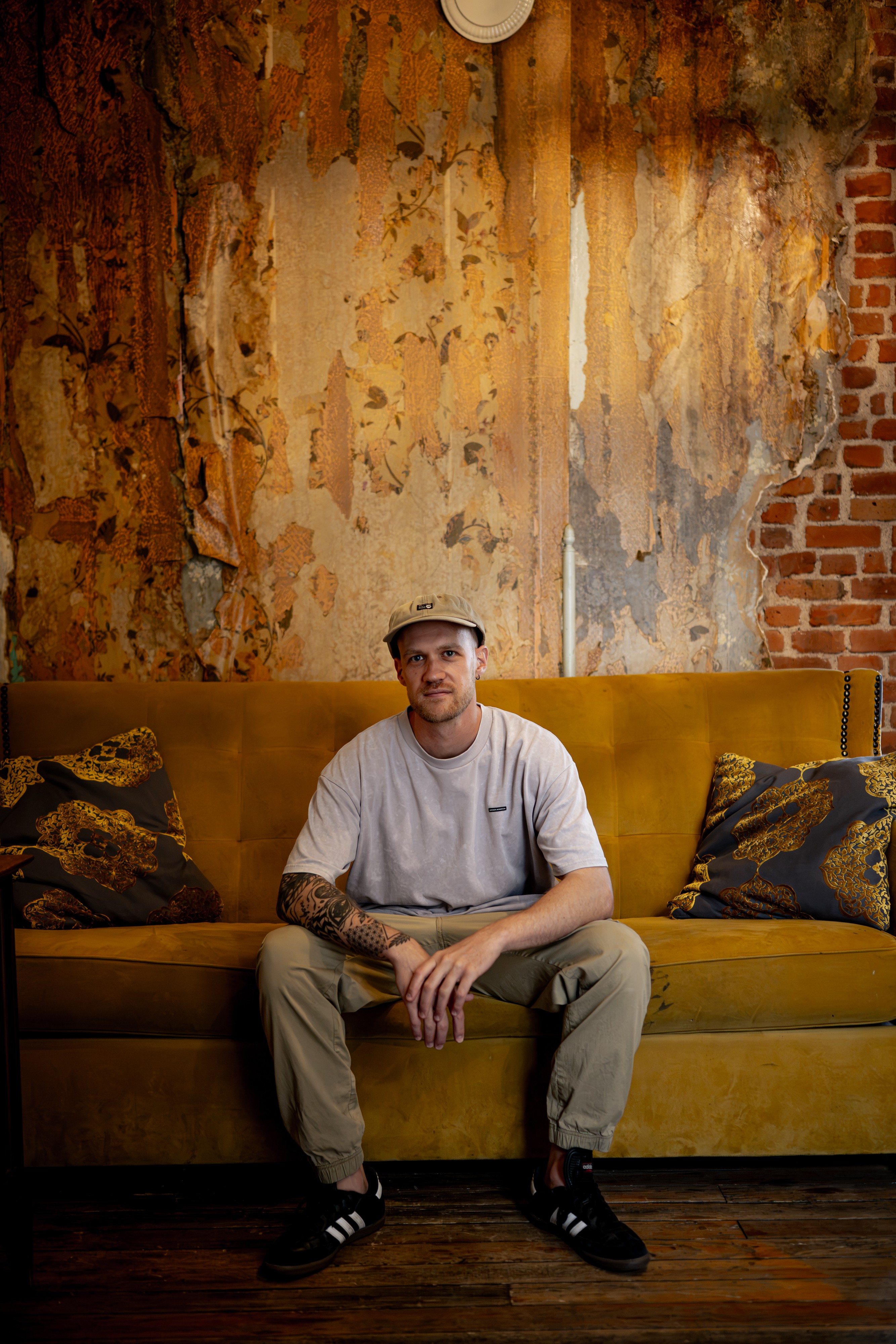 Man sitting on a yellow sofa with pillows, in front of a textured wall with peeling paint and exposed brick.