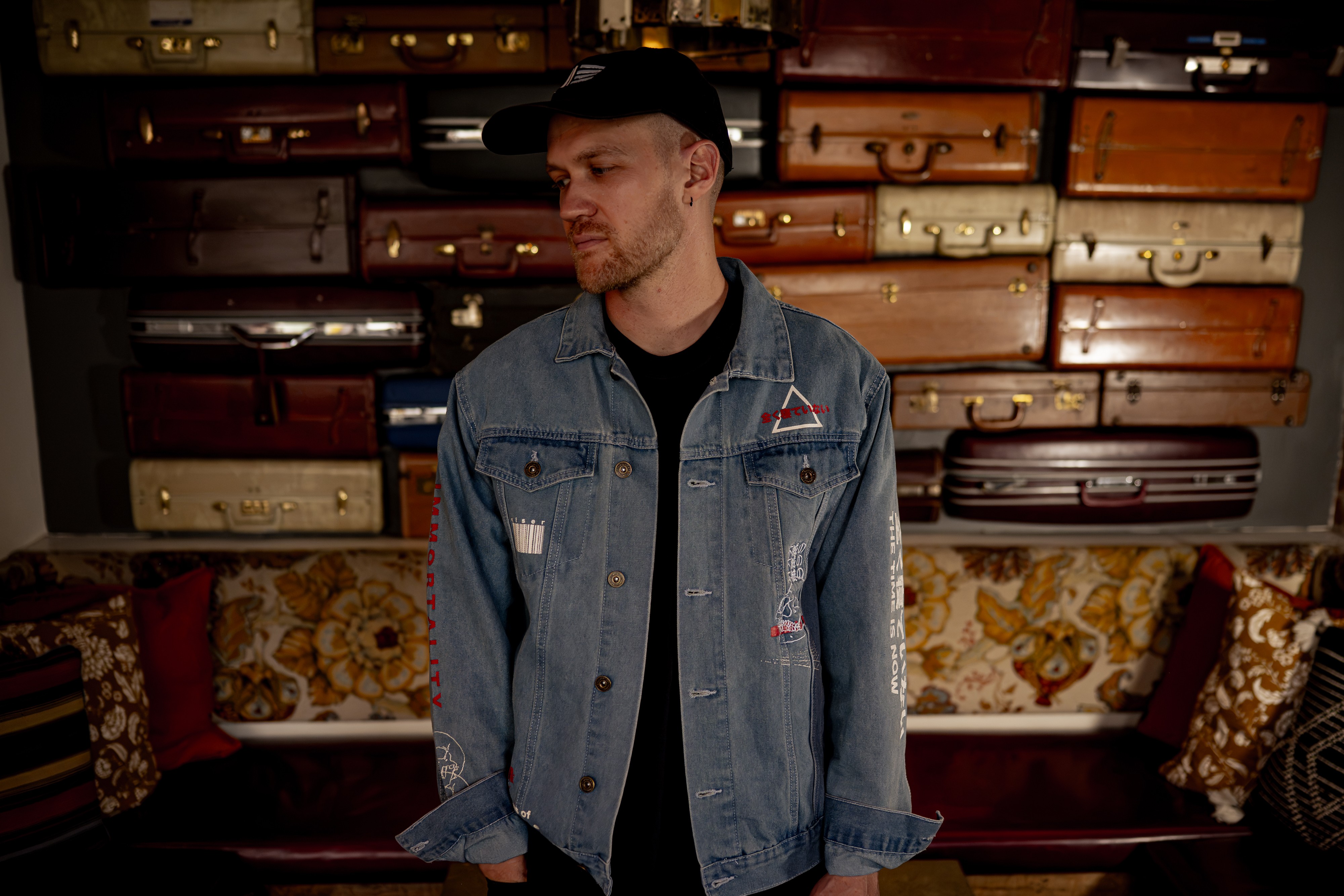 Man in denim jacket and black cap sitting on a vintage sofa with patterned cushions, shelves with suitcases behind him.