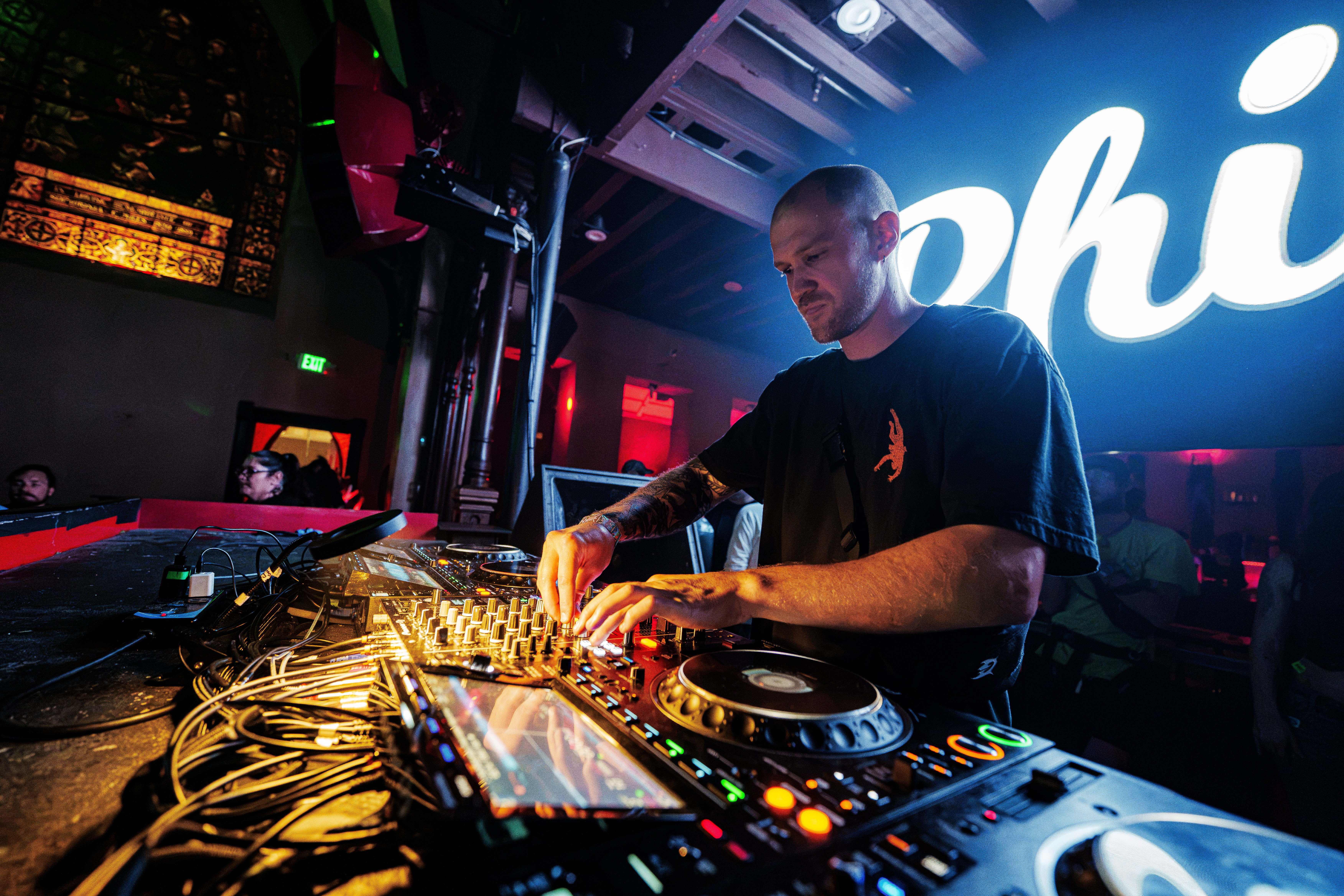 DJ mixes music at a club with a large illuminated sign in the background, colorful lights, and equipment on a table.