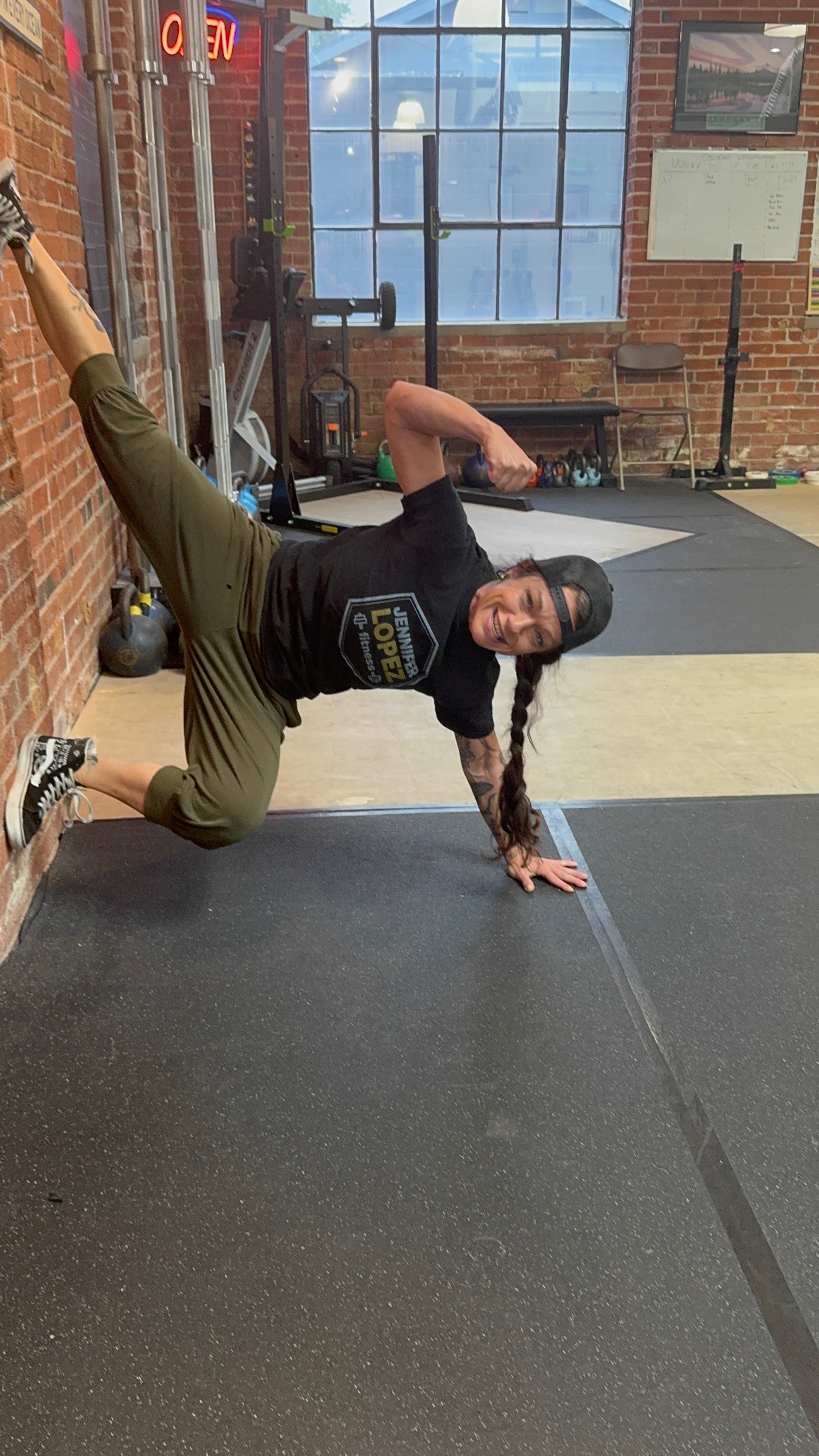 Woman in black shirt and green pants performs a side plank exercise on a gym floor, with brick walls and windows in background.