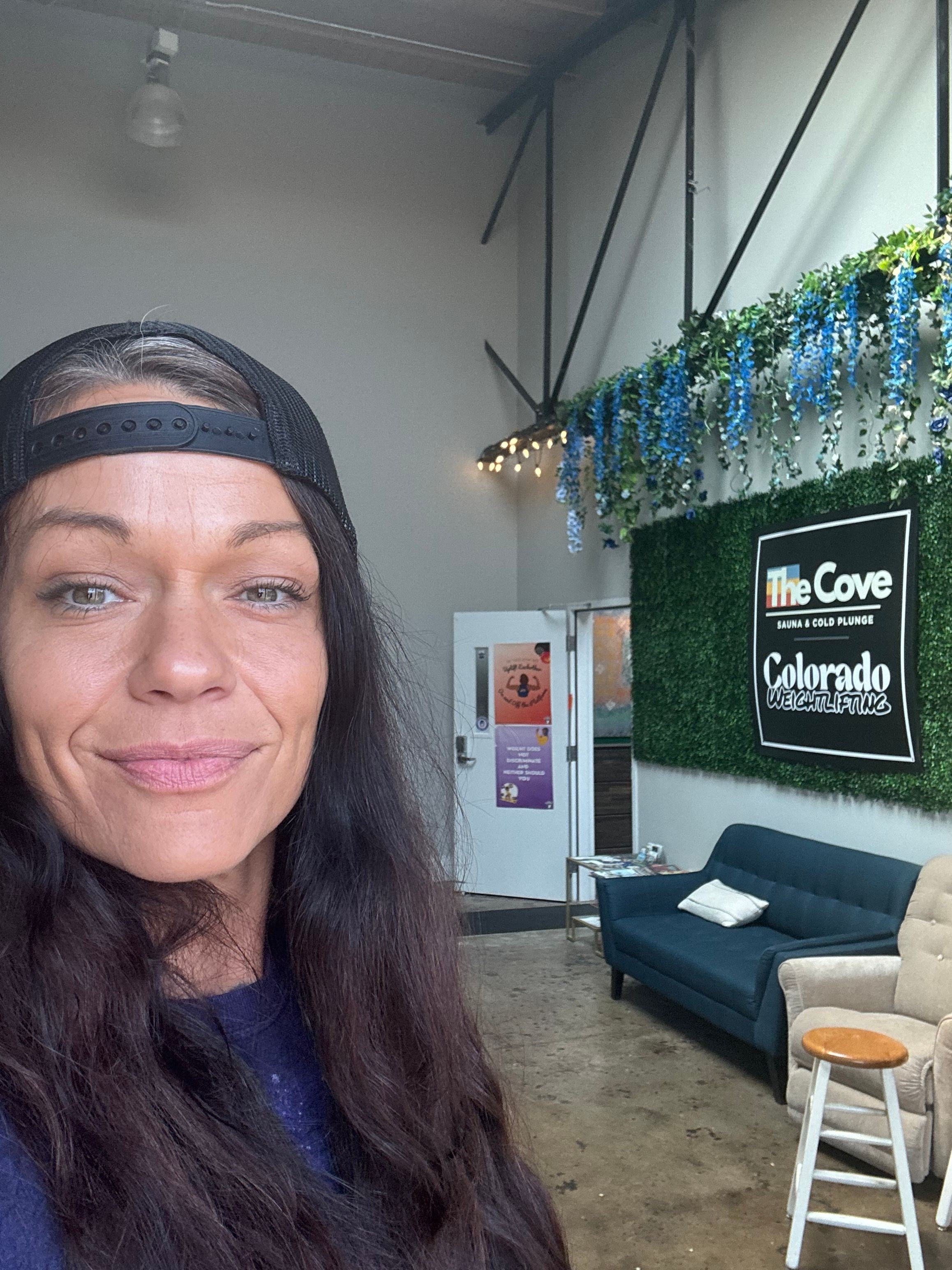Woman with long dark hair and a headband, smiling indoors with a decorated wall and seating area behind her.