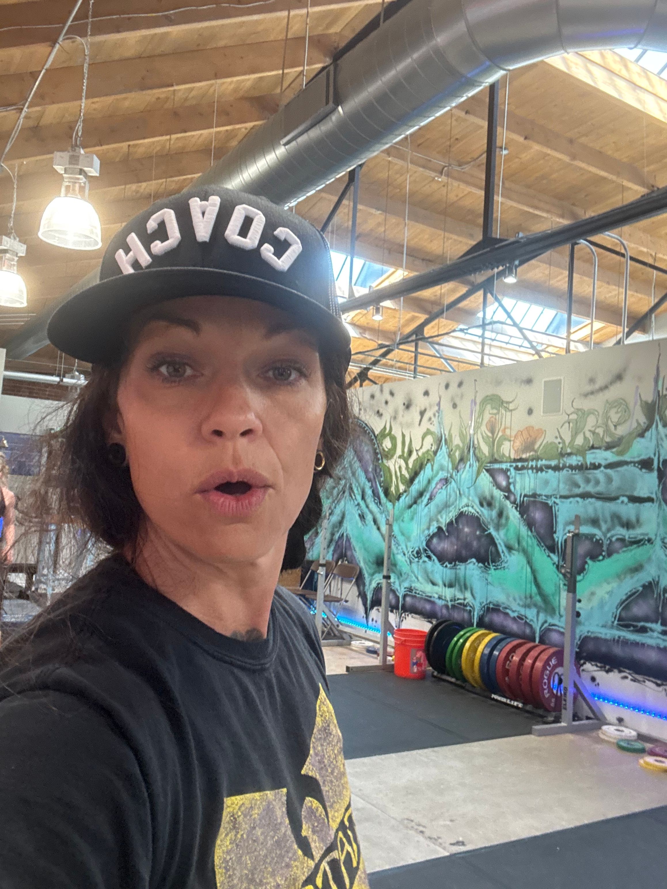 Woman wearing a black cap with 'COACH' written on it, taking a selfie indoors with colorful artwork in the background.