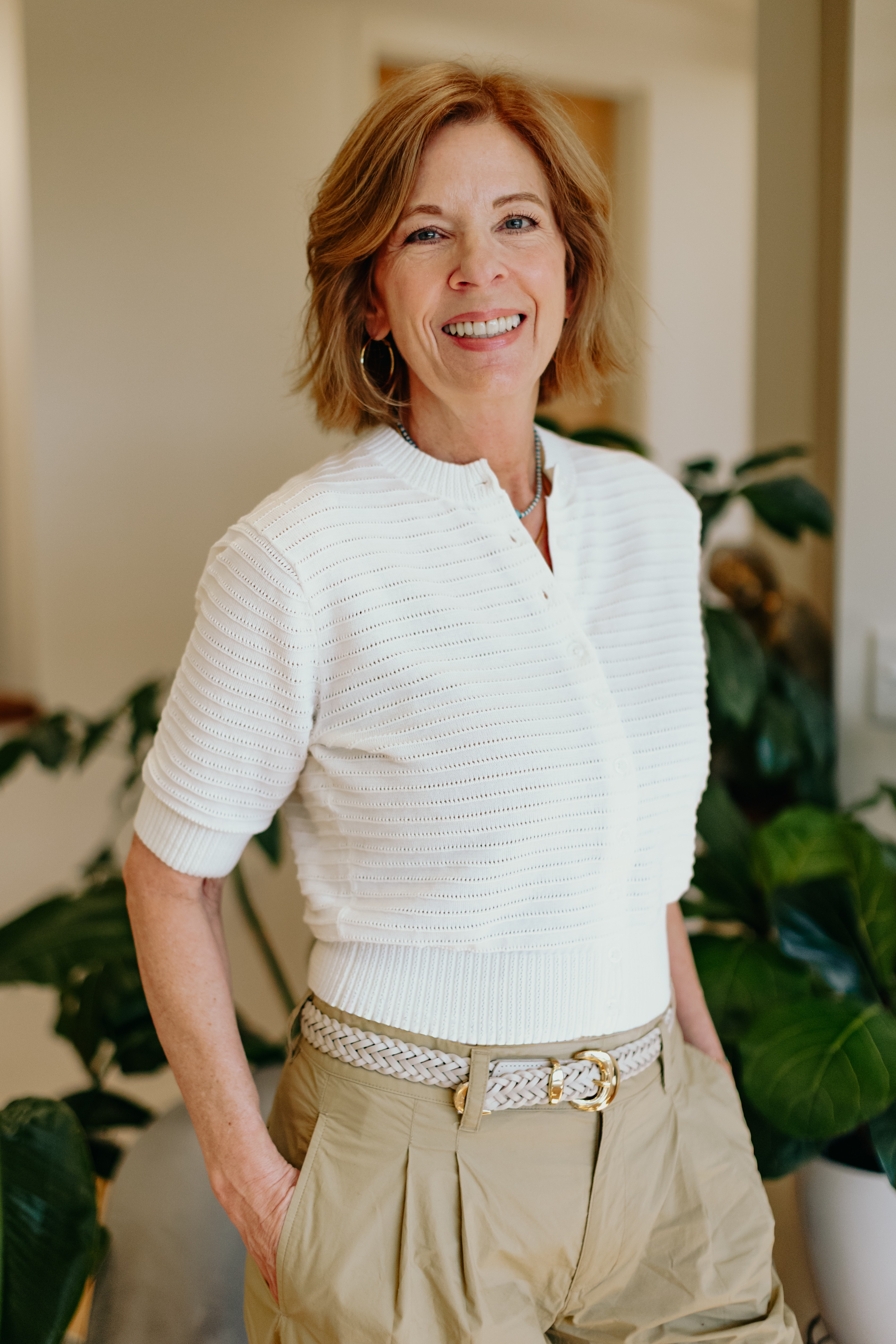 Smiling woman with short red hair wearing a white top and beige pants, standing indoors near plants.