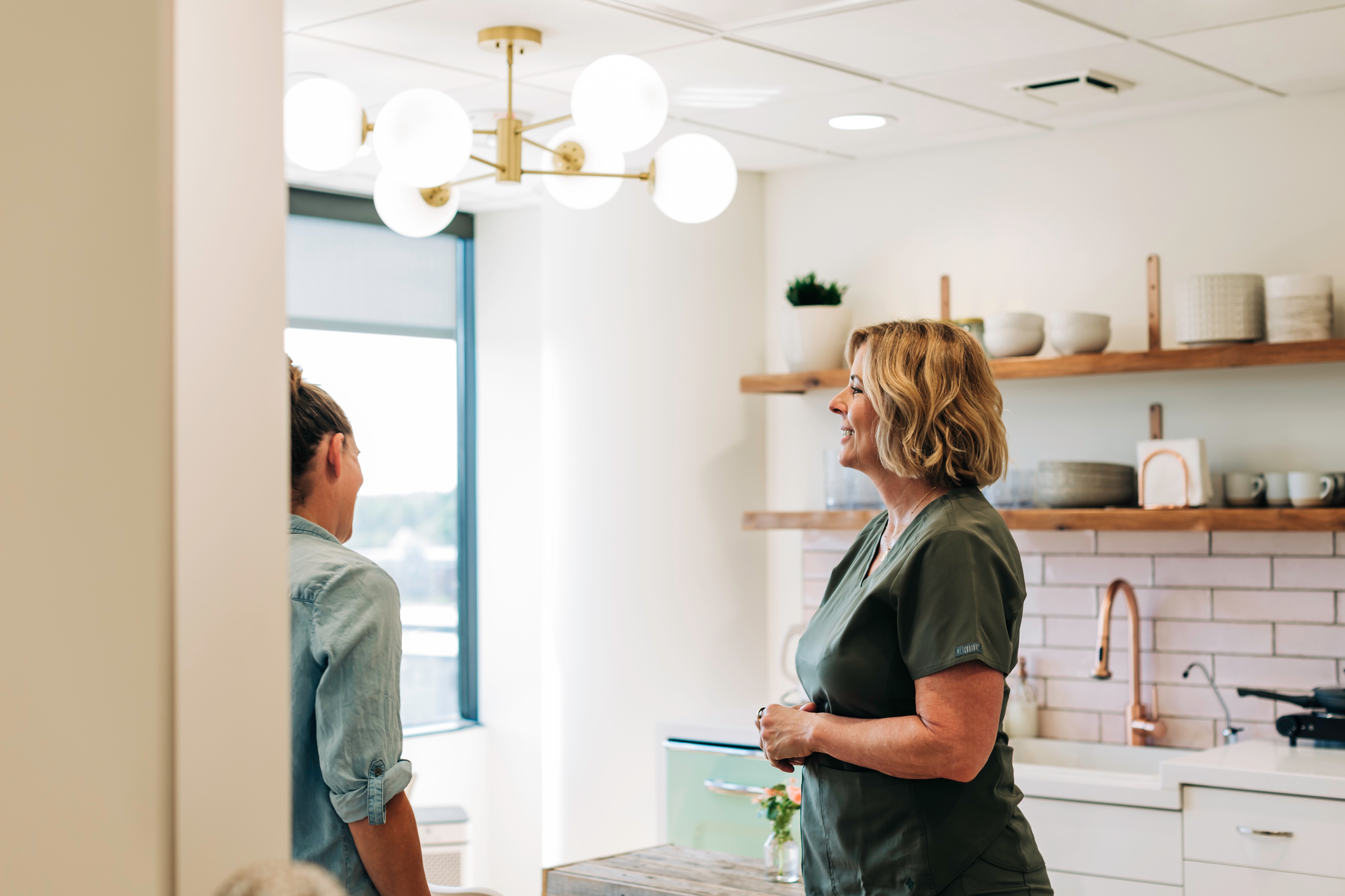 Two women in a modern kitchen with white walls, wooden shelves, and a window, engaging in conversation.