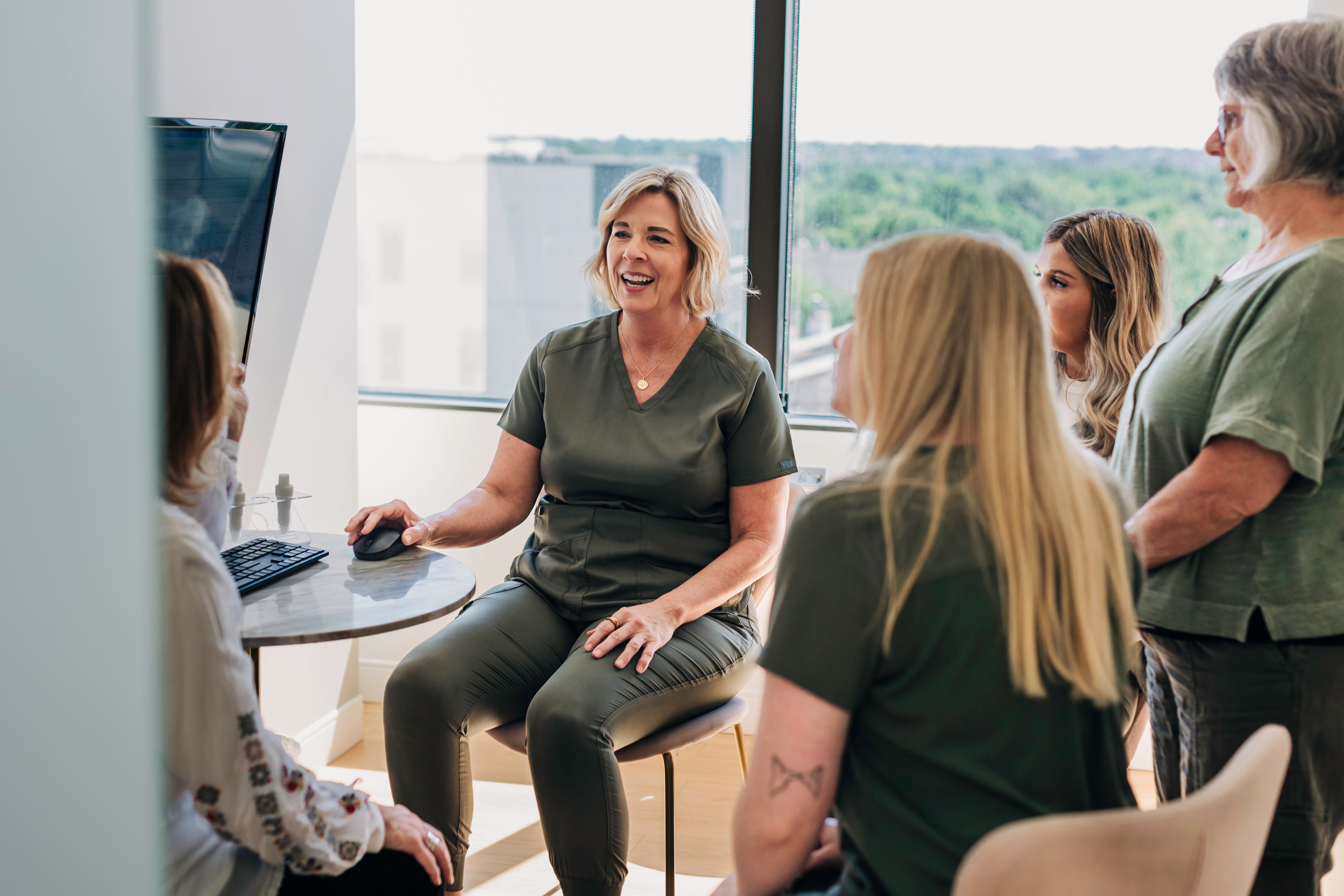 Group of five women in a meeting room, one woman speaking and others listening, with a large window in the background.