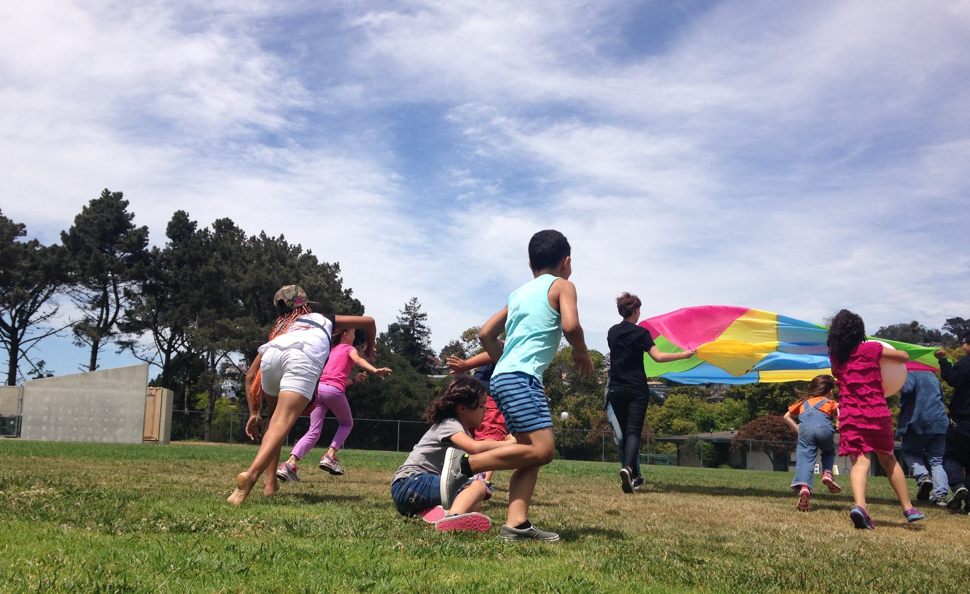 Children playing outdoors on grass with a colorful parachute, trees, and a partly cloudy sky in the background.