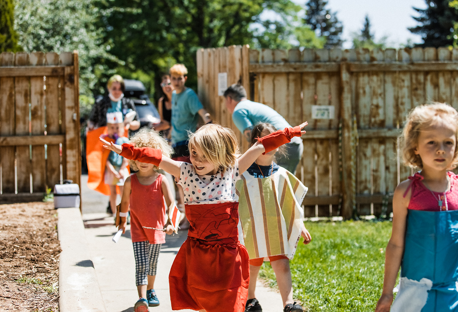 Children playing outdoors in a backyard with a wooden fence, some holding water guns, sunny day, trees in background.
