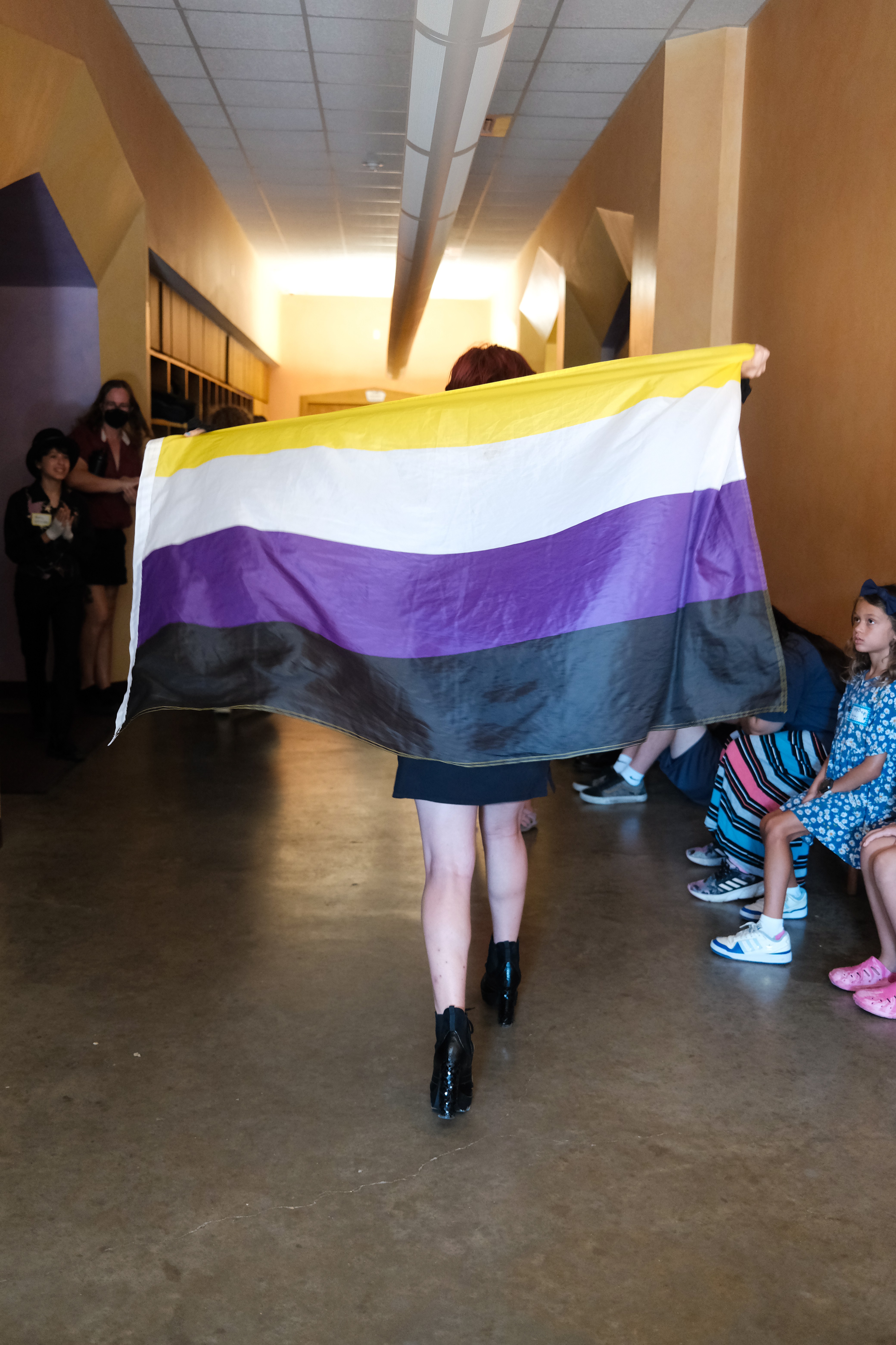 Person holding a rainbow pride flag walking in a hallway with people seated on the side, walls painted yellow and brown.