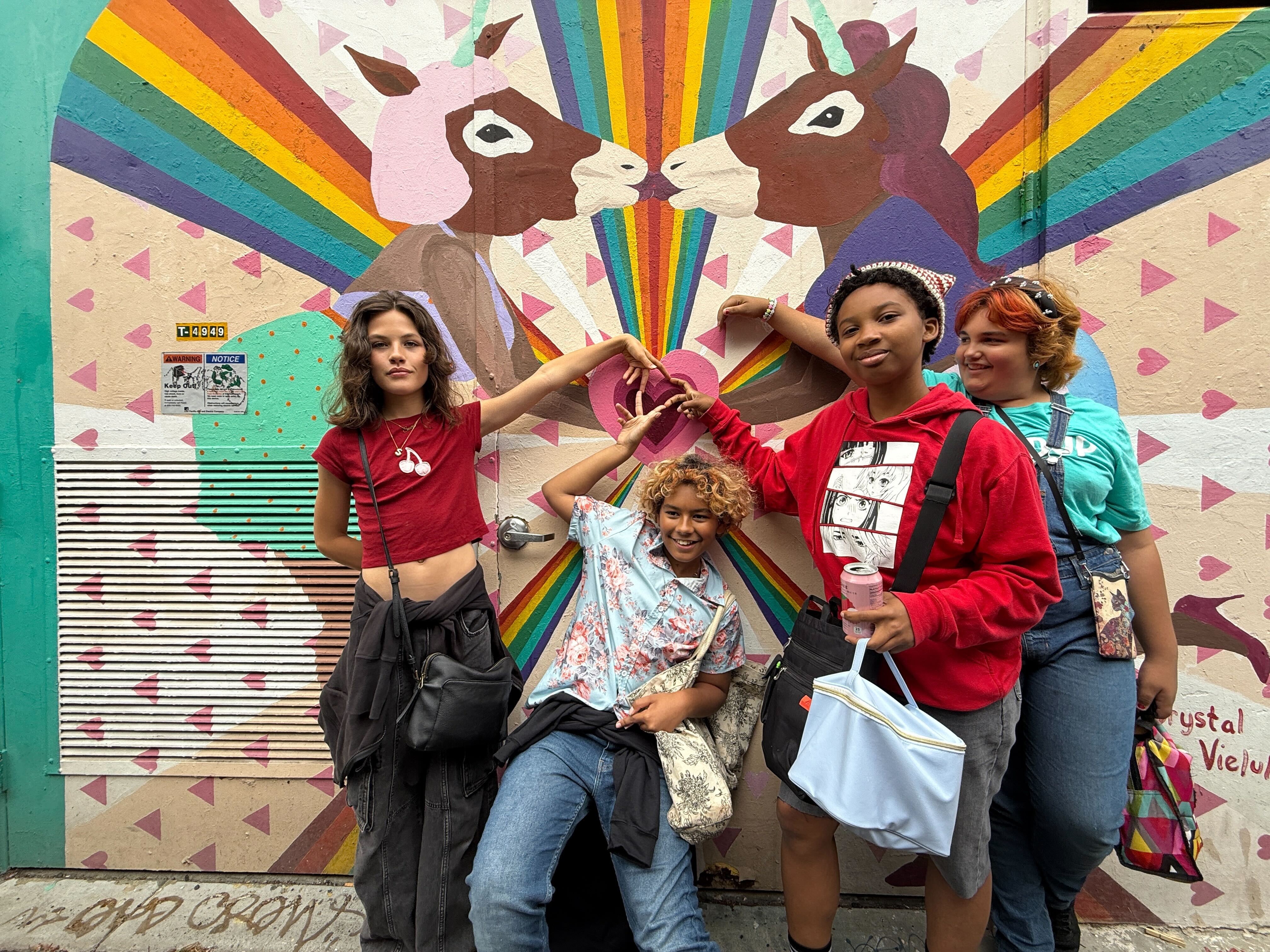 Four young women stand in front of a colorful mural with two unicorns and rainbow rays, holding hands and smiling.