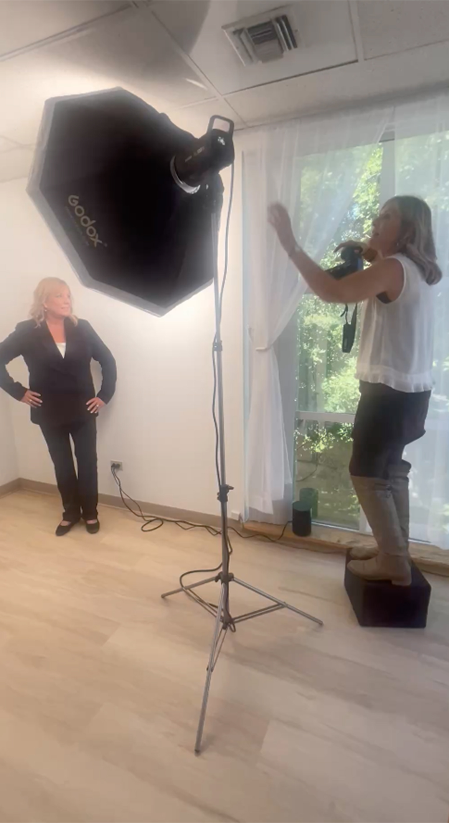 Two women in a room with a large softbox light, one gesturing with hands, the other standing with hands on hips, window in background.