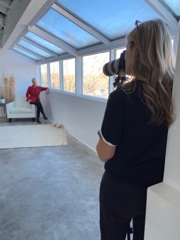 Person with long hair taking a photo of another person sitting near windows in a bright room.
