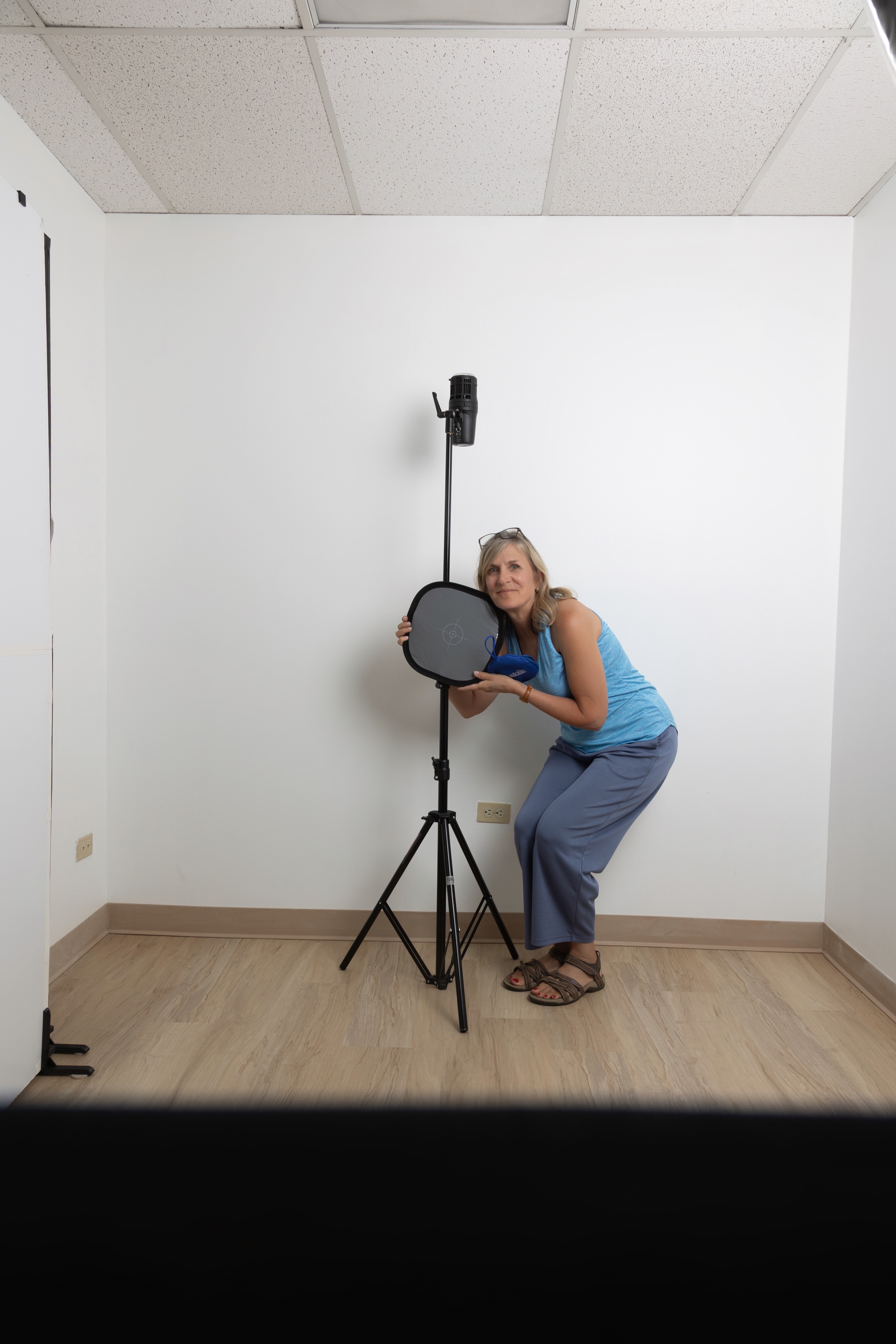 Woman in blue top and pants crouching next to a camera on a tripod in a plain white room with a wooden floor.