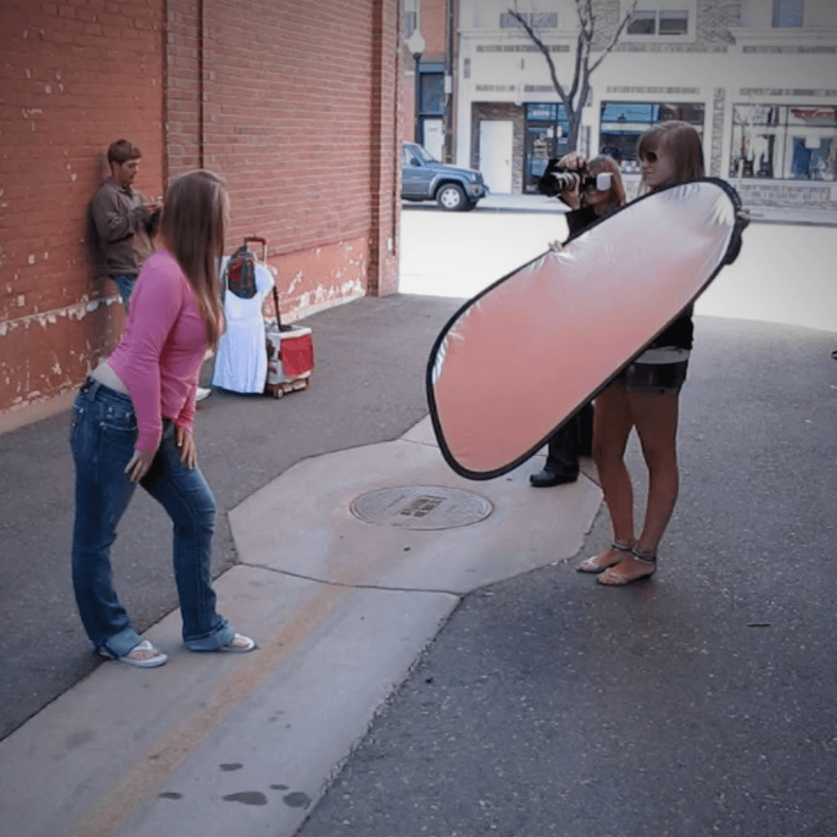 Photographer takes a picture of a woman against a brick wall with a reflector, in an urban setting.