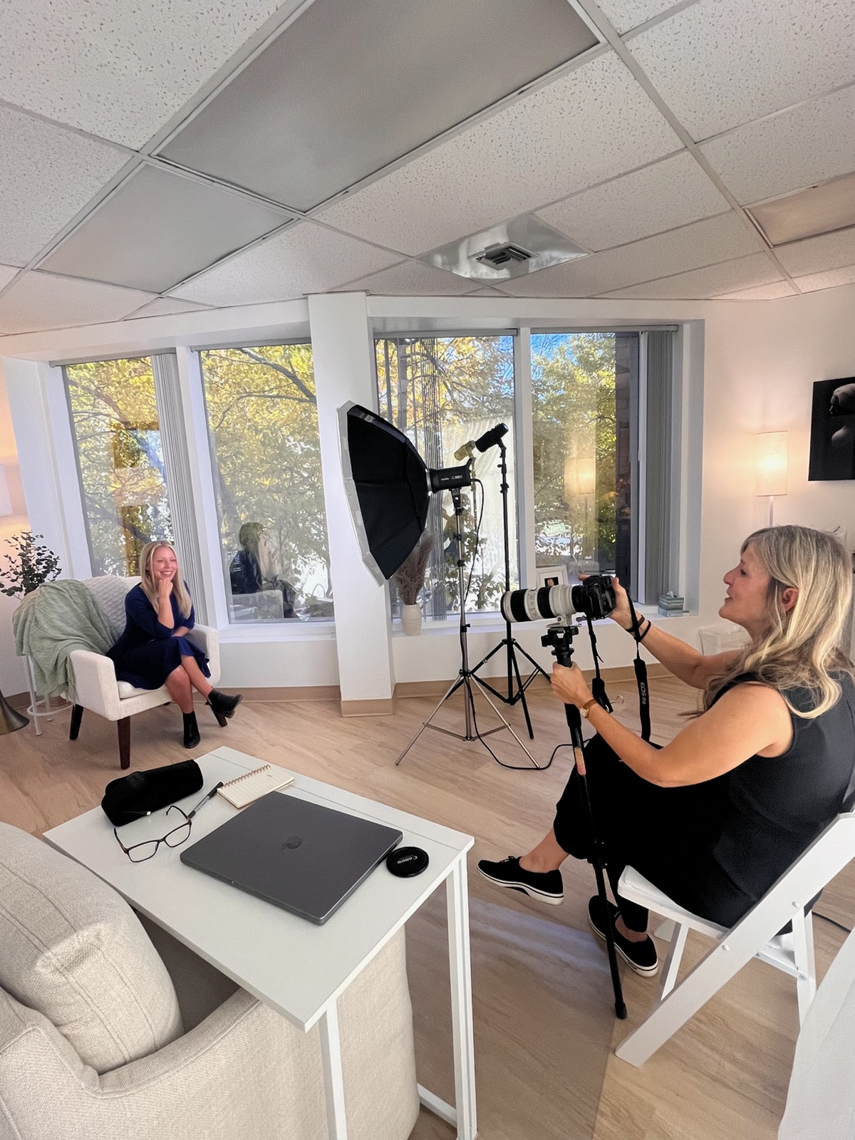 Woman sitting in a chair being photographed by a photographer in a bright room with large windows and studio lighting.